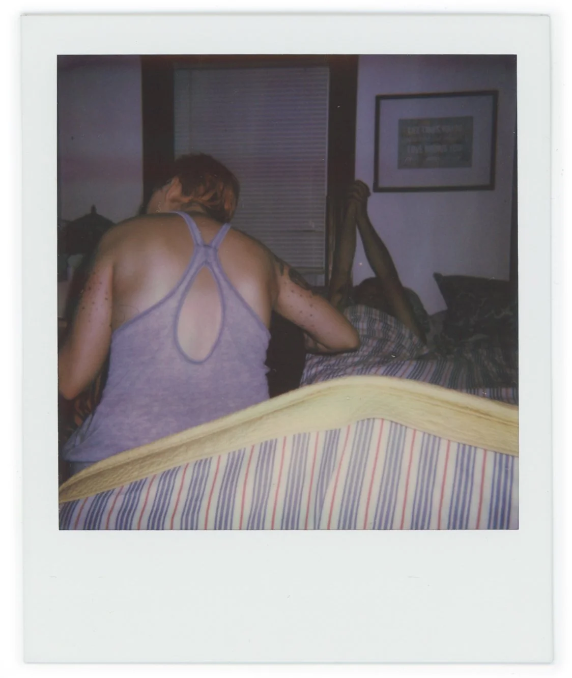 A woman with short reddish hair in a purple tank top is kneeling on the bed, facing away from the camera, next to a person lying in bed. The person in bed is holding their legs up with their hands. The room has a window with closed blinds and framed 