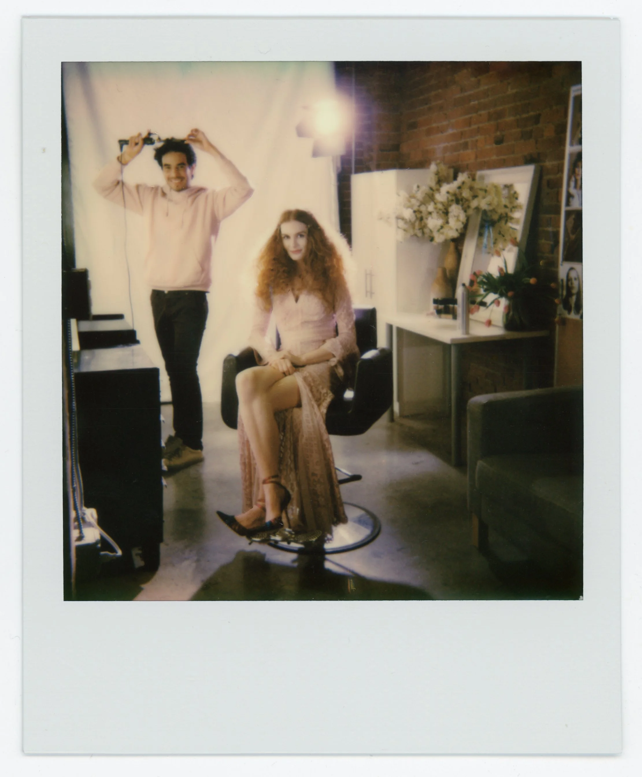 A woman with curly red hair and a pink lace dress sitting in a hairstylist chair, holding a hairbrush, with a man standing behind her holding a hairdryer. The setting appears to be a salon or photo studio with a brick wall, flowers, and photography e