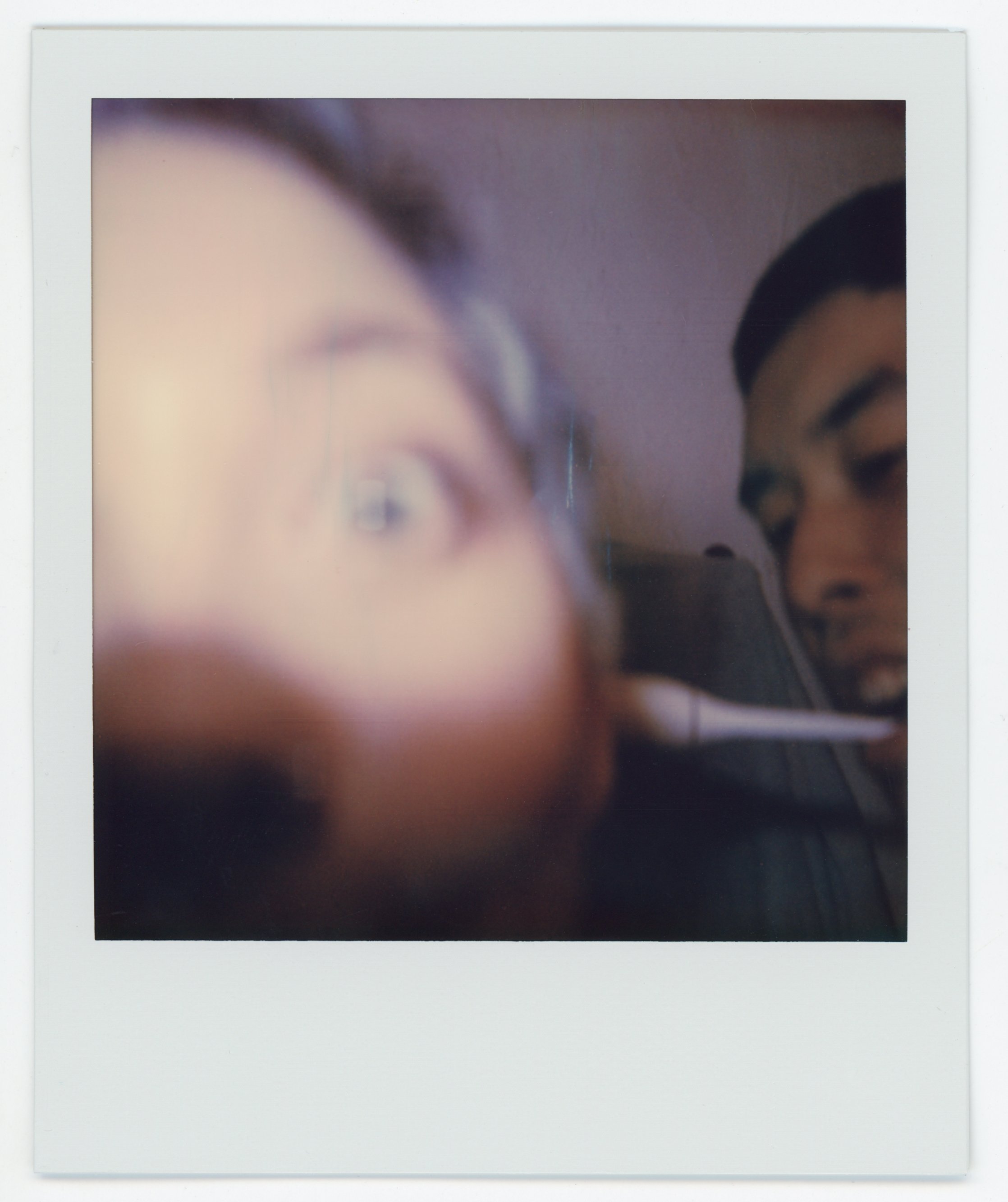 A blurry Polaroid photo showing two people, one in the foreground with glasses and another in the background, both engaged with a smartphone.