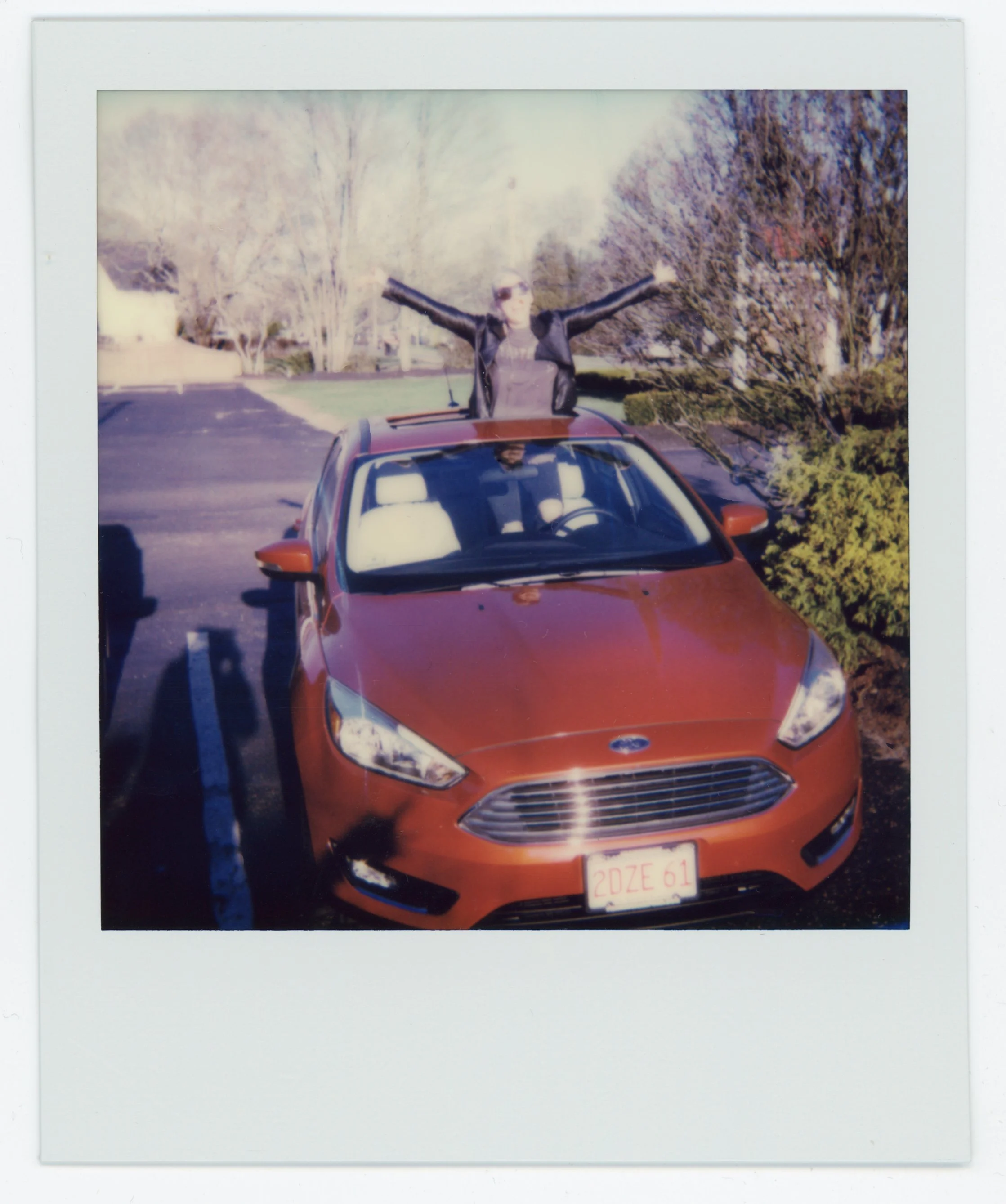 Person standing on a red car with arms outstretched in a parking lot, trees and bushes in the background, sunny day, Polaroid photo