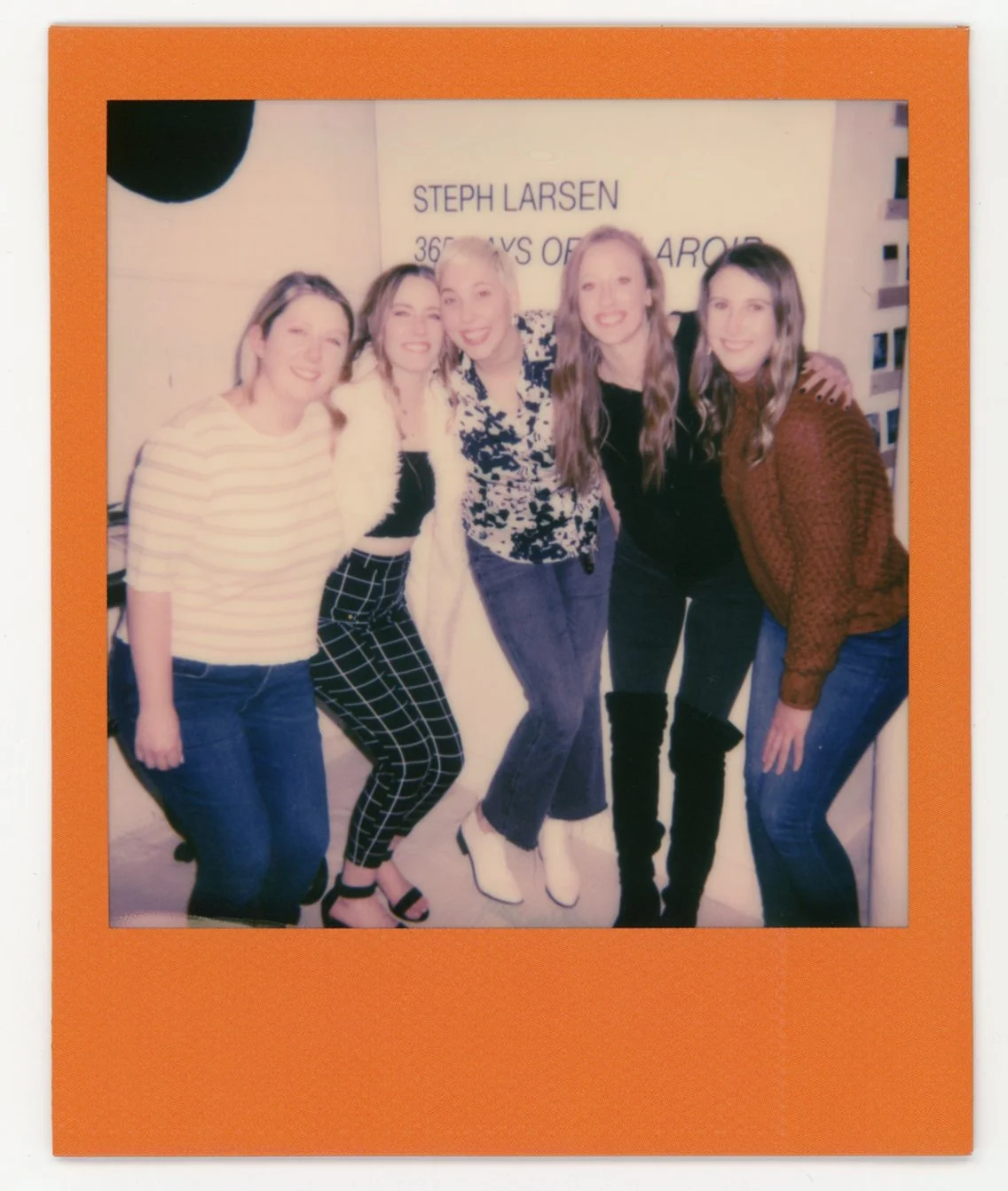 Five women smiling and posing together at an indoor event, with a sign and shelf in the background.