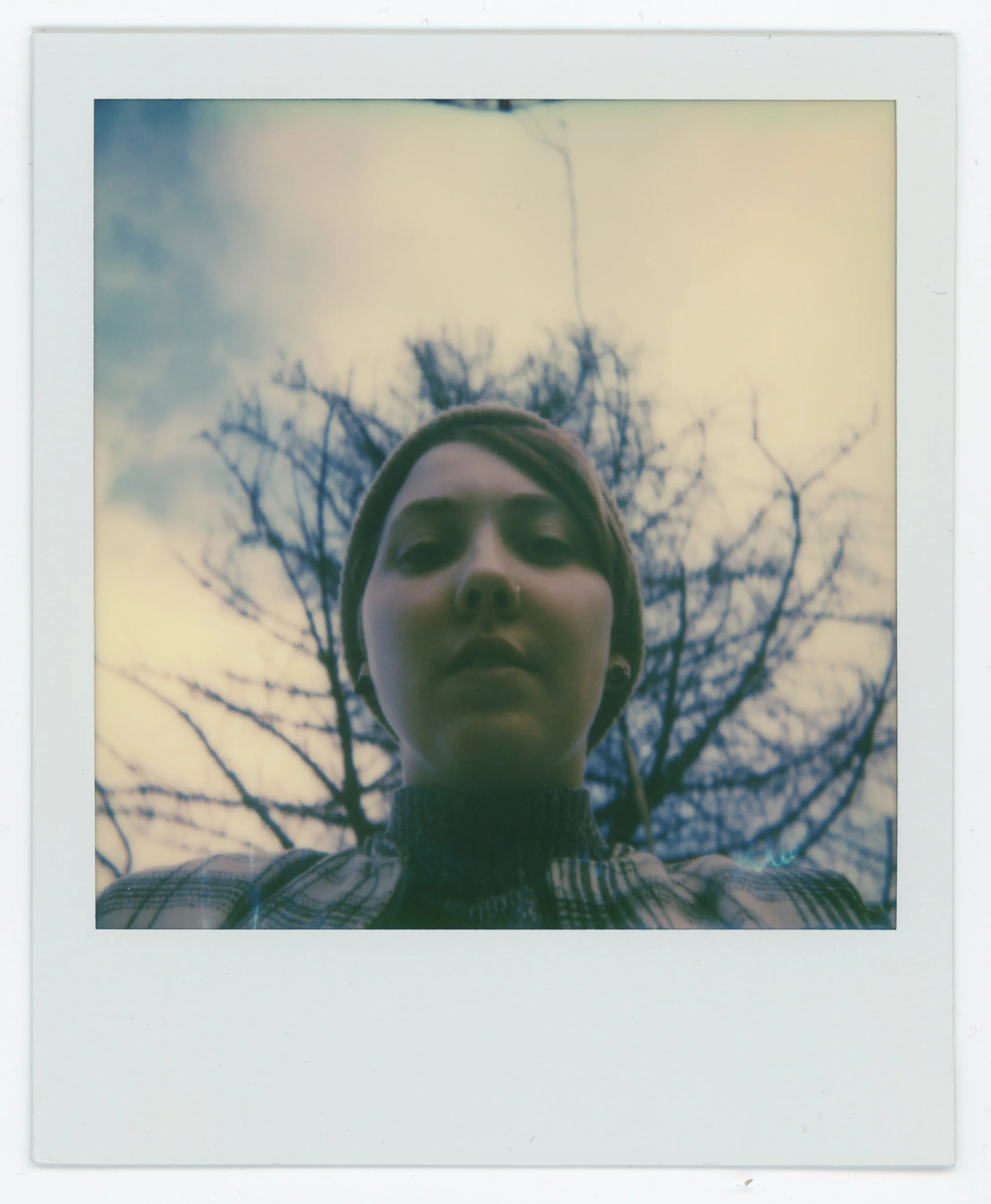 A close-up selfie of a woman with short hair, wearing a beanie and a plaid coat, taken outdoors during the daytime. Behind her, a leafless tree and cloudy sky are visible.