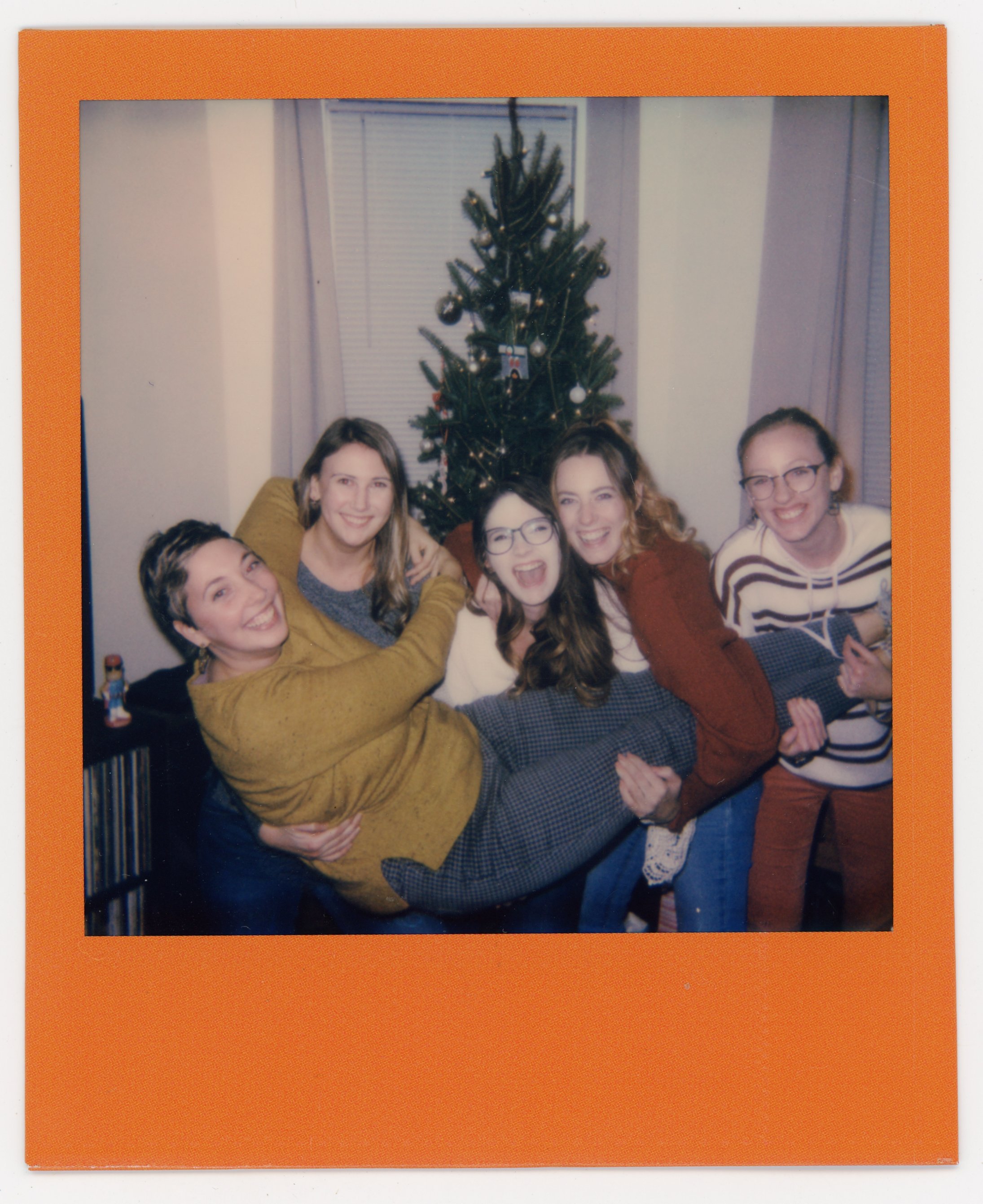 Group of five young women smiling and holding one woman horizontally in front of a decorated Christmas tree in a living room.