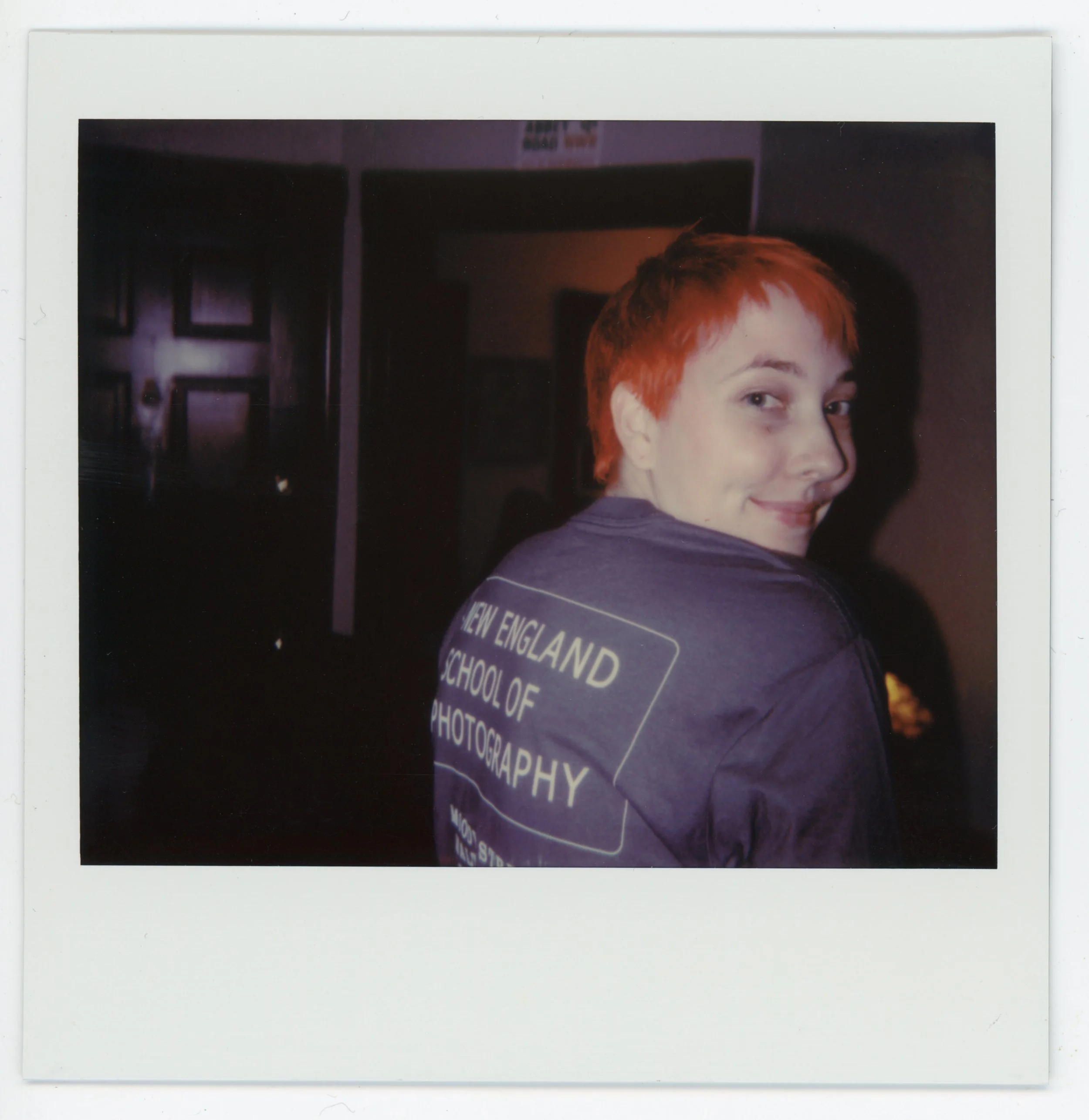A person with short, red hair smiling and turning their head to the side, wearing a blue sweatshirt with white text on the back that reads "NEW ENGLAND SCHOOL OF PHOTOGRAPHY."