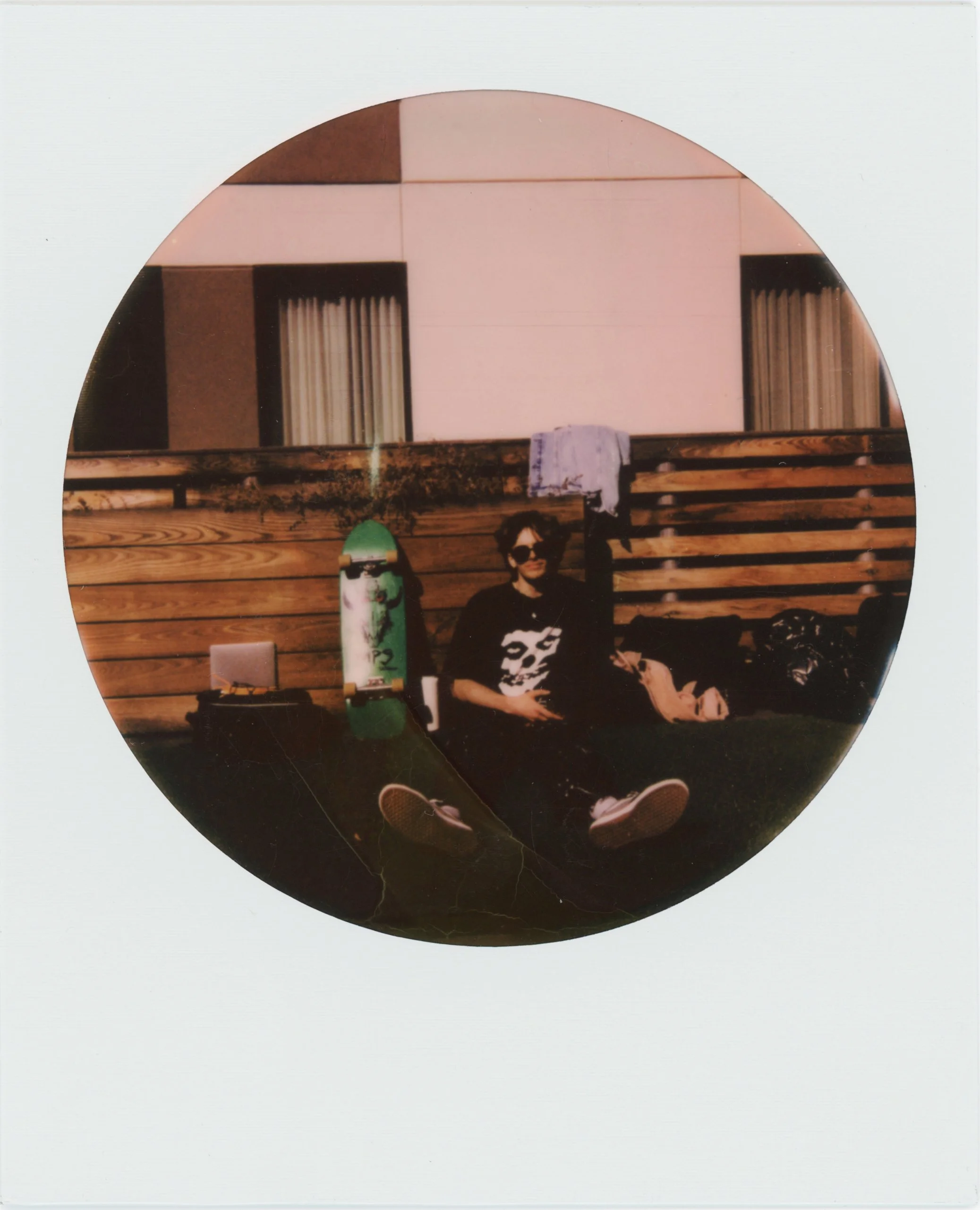 A person with sunglasses and a skull shirt sitting on grass against a wooden fence, skateboard leaning against the fence, and various belongings nearby, with a building in the background.