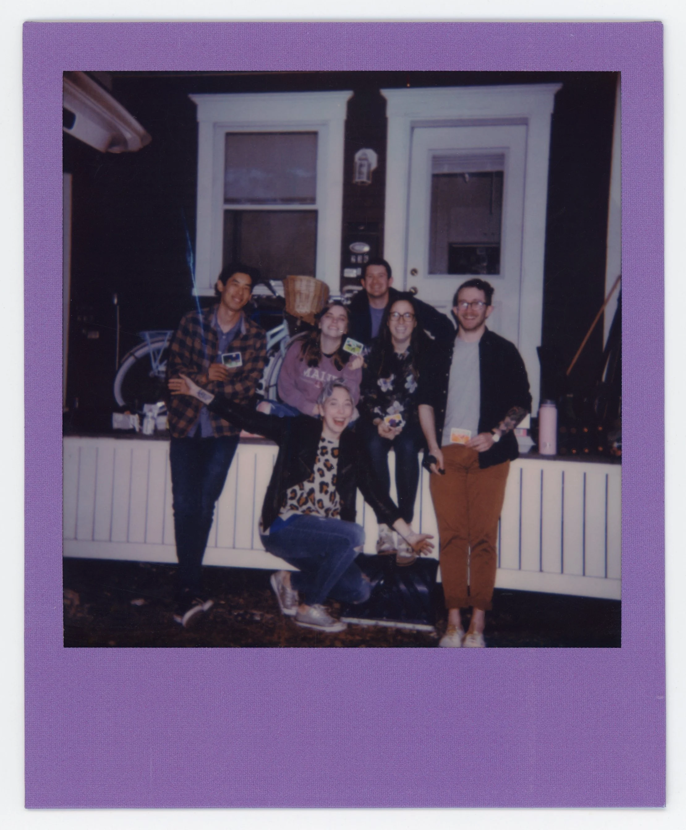Group of six young adults posing outside a house, some holding photographs, smiling and celebrating.