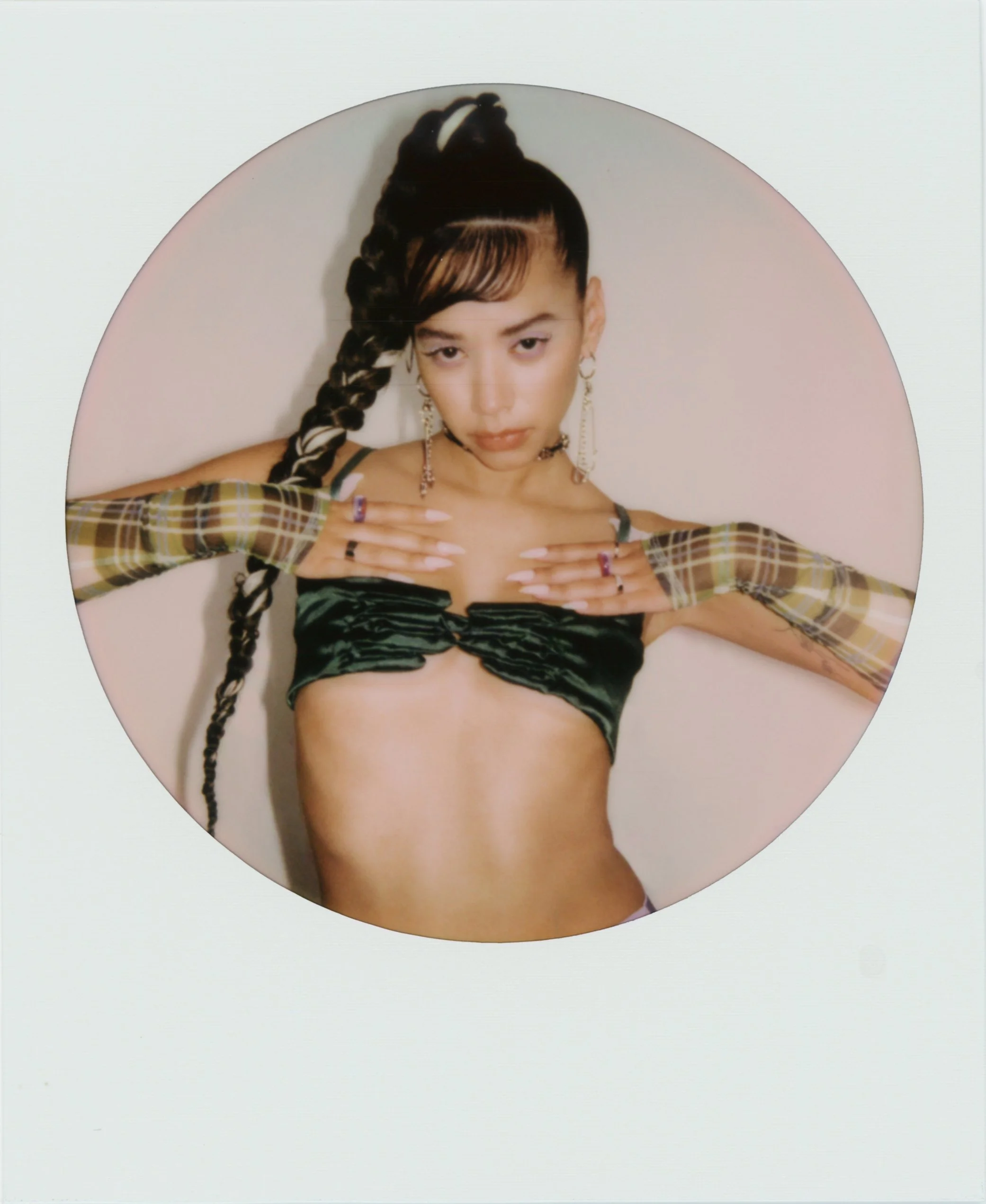 A woman with long braided hair, wearing a black velvet crop top, patterned gloves, and large earrings, laying on a pink surface with a white background.