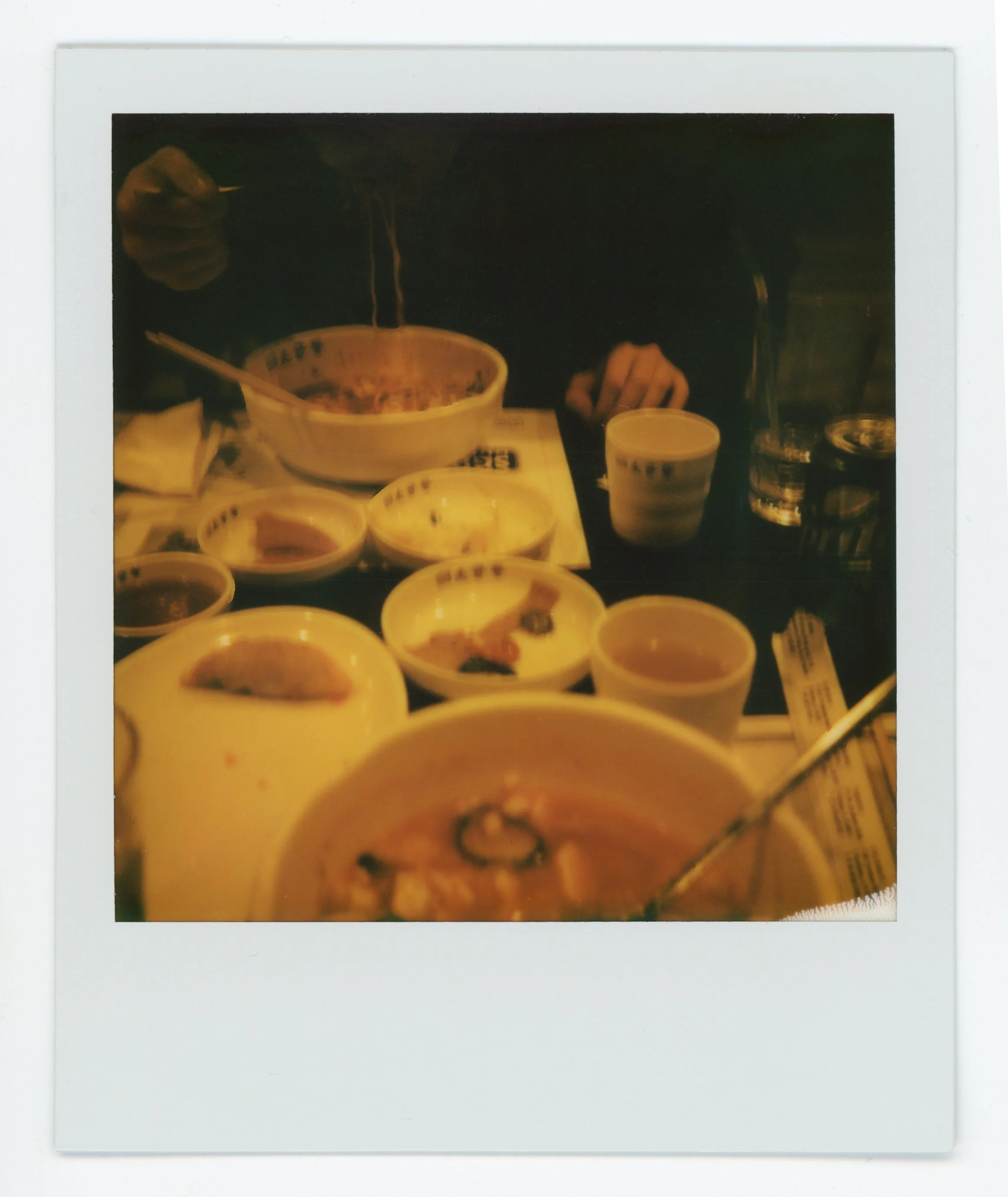 Polaroid photo of a table filled with bowls of ramen, bowls of dipping sauces, a glass of water, and a person holding a cup, with chopsticks in hand.