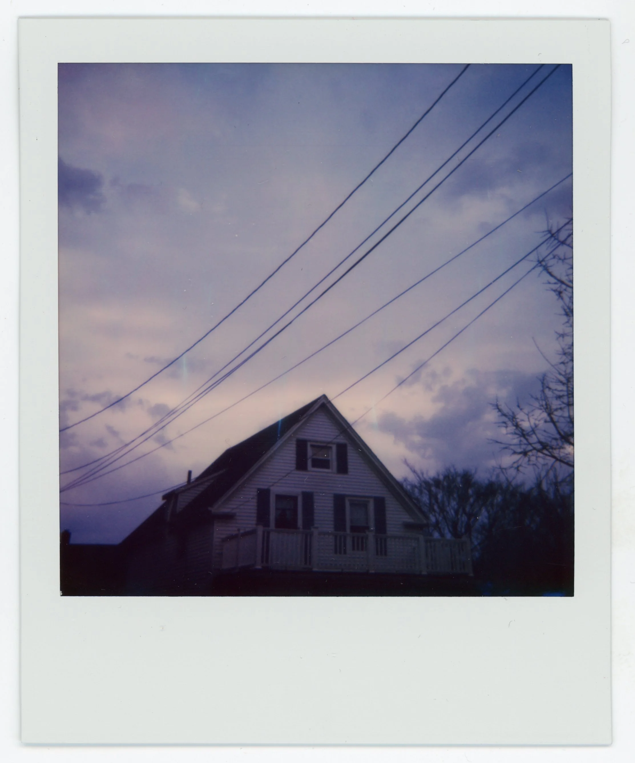 A house with a peaked roof and a balcony, set against a cloudy sky with power lines overhead.