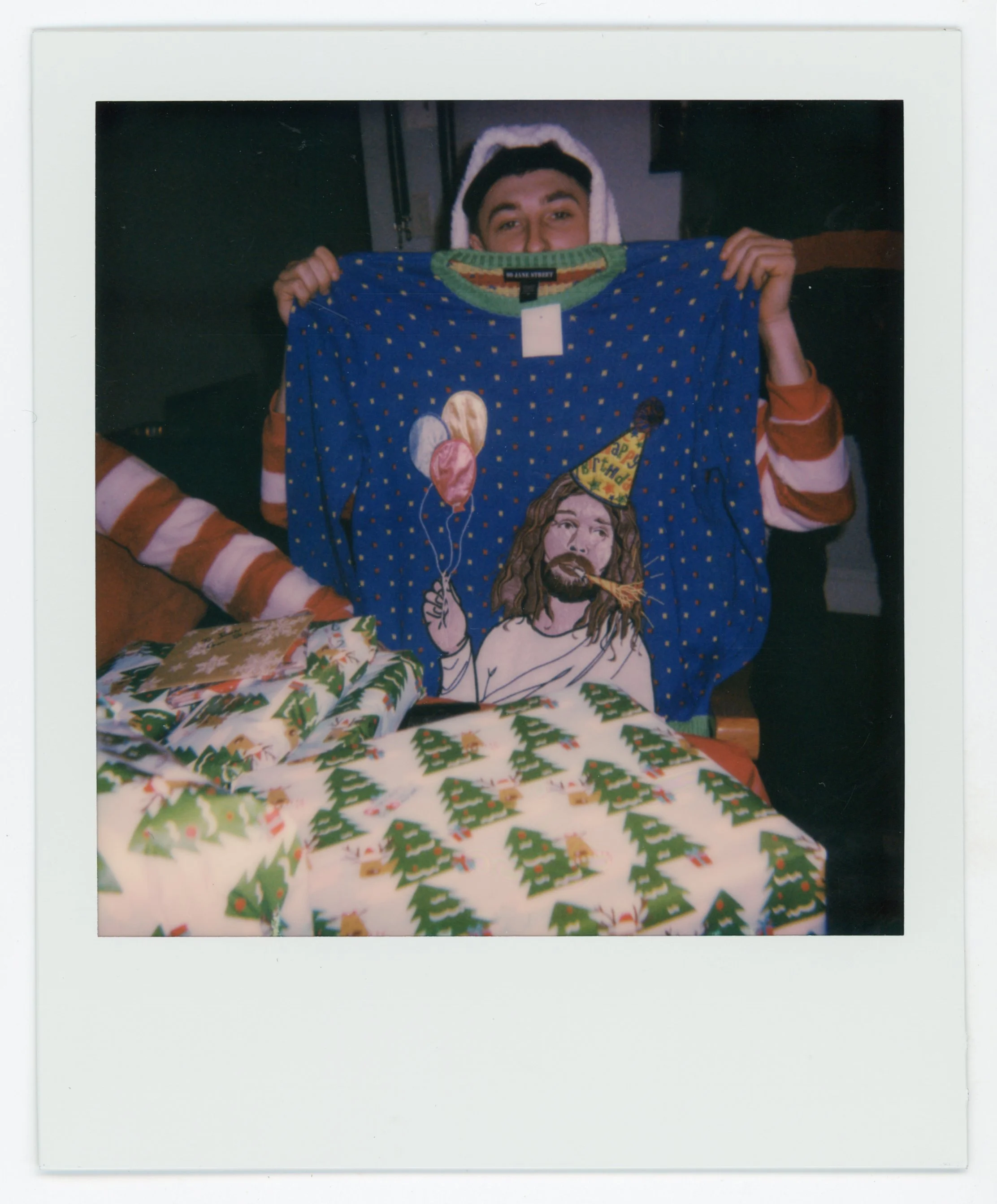 Person holding up a blue Christmas sweater with a Jesus graphic, surrounded by wrapped Christmas presents.