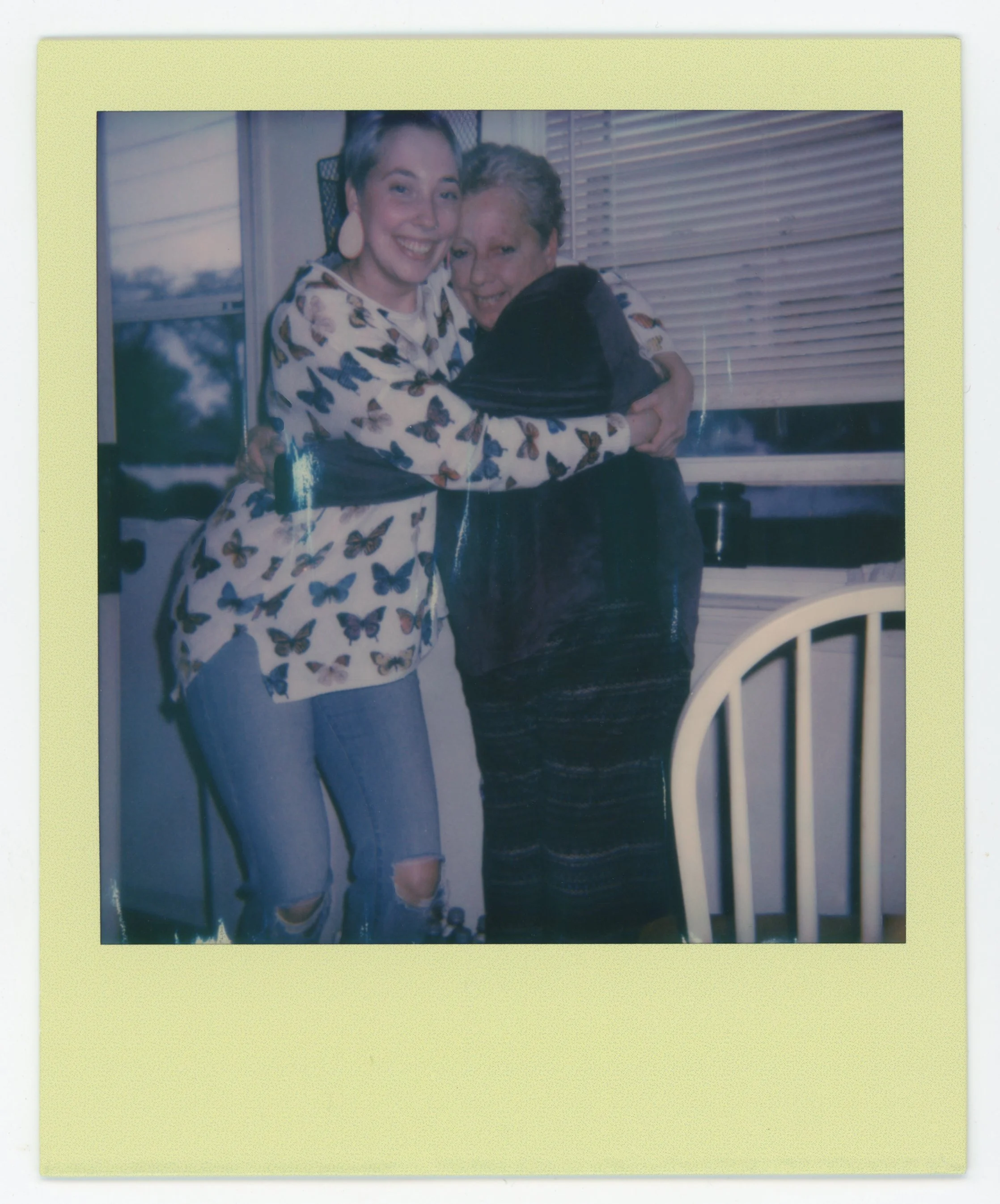 Two women hugging inside a kitchen near a window, smiling, one wearing a butterfly print shirt and ripped jeans, the other in a black jacket and striped pants.