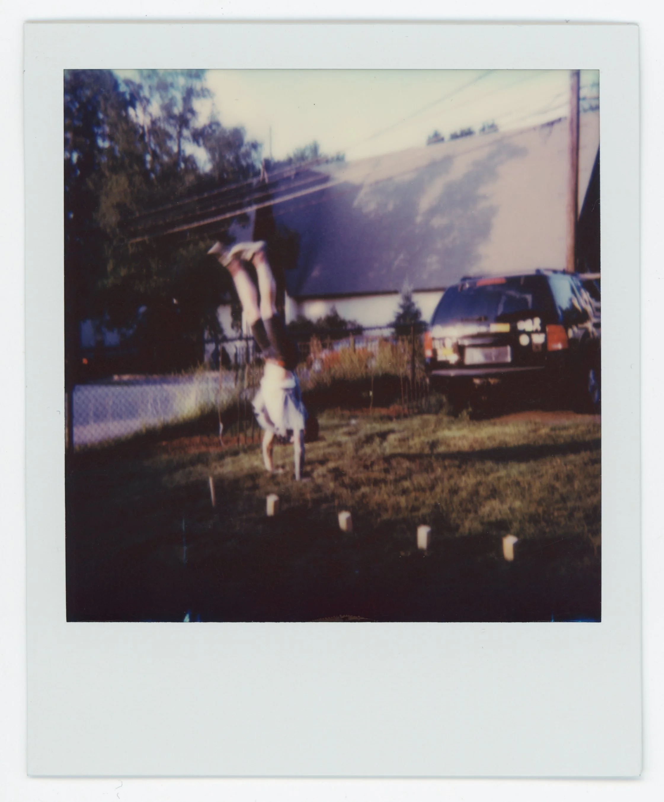 A blurry Polaroid photo showing a person performing a handstand outdoors near a house and a parked car.