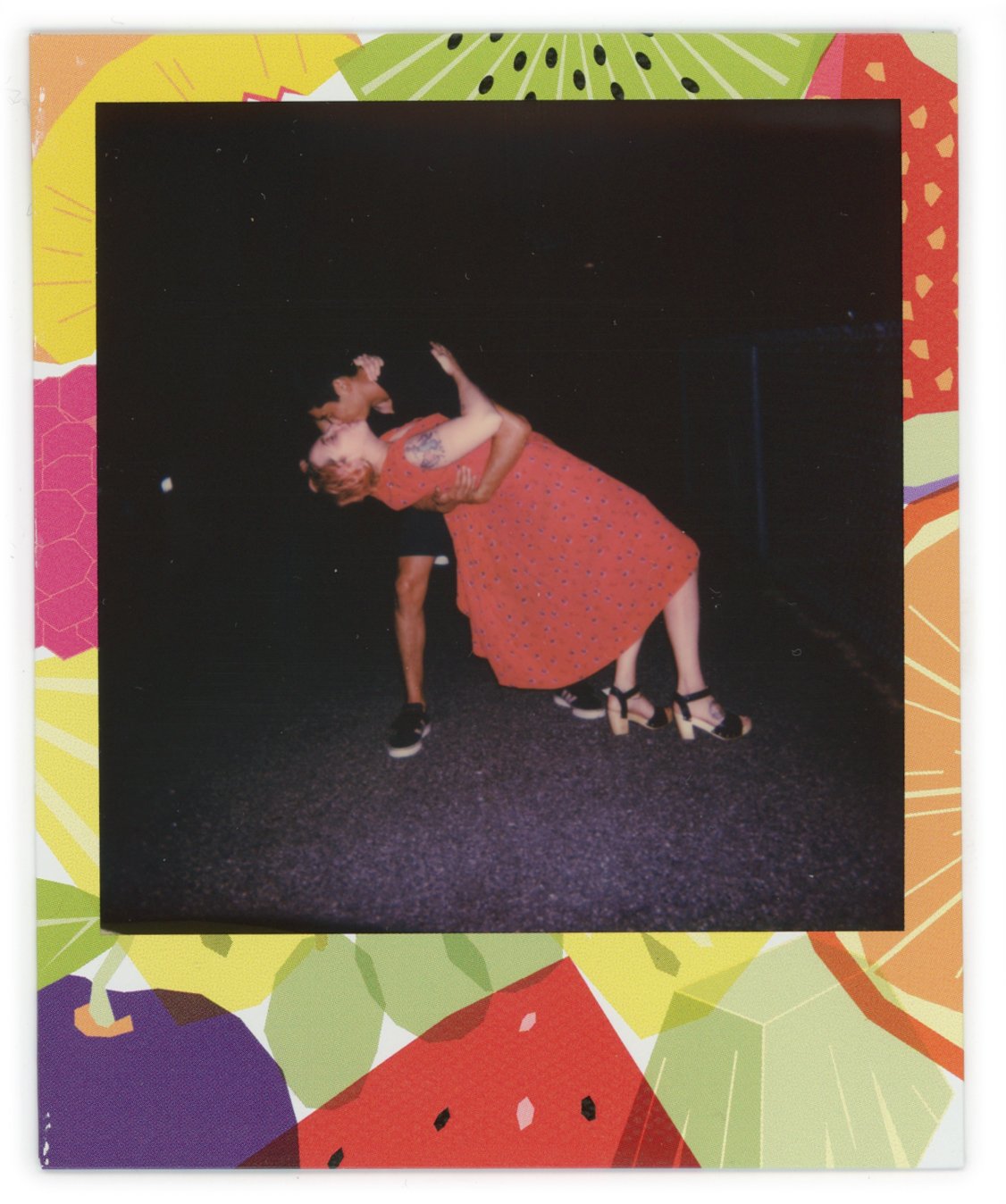 Two people dancing closely on a dark background, one person wearing a red dress with small blue dots and the other person leaning back, wearing black shoes. The photo is framed with a colorful fruit-patterned border.