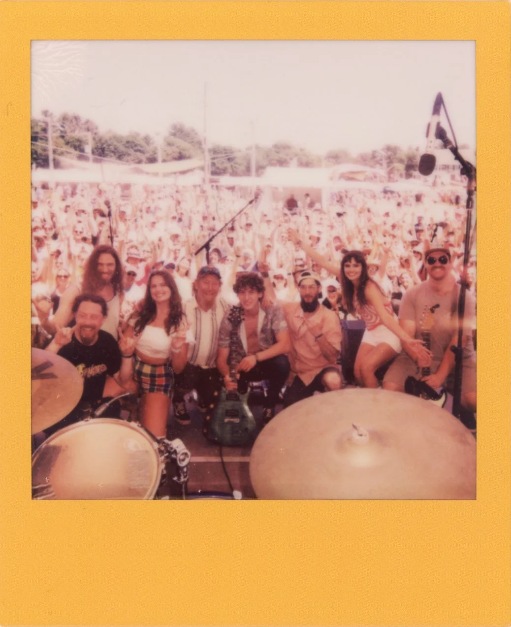 A group of musicians and friends posing on stage at an outdoor music festival with a large crowd in the background.