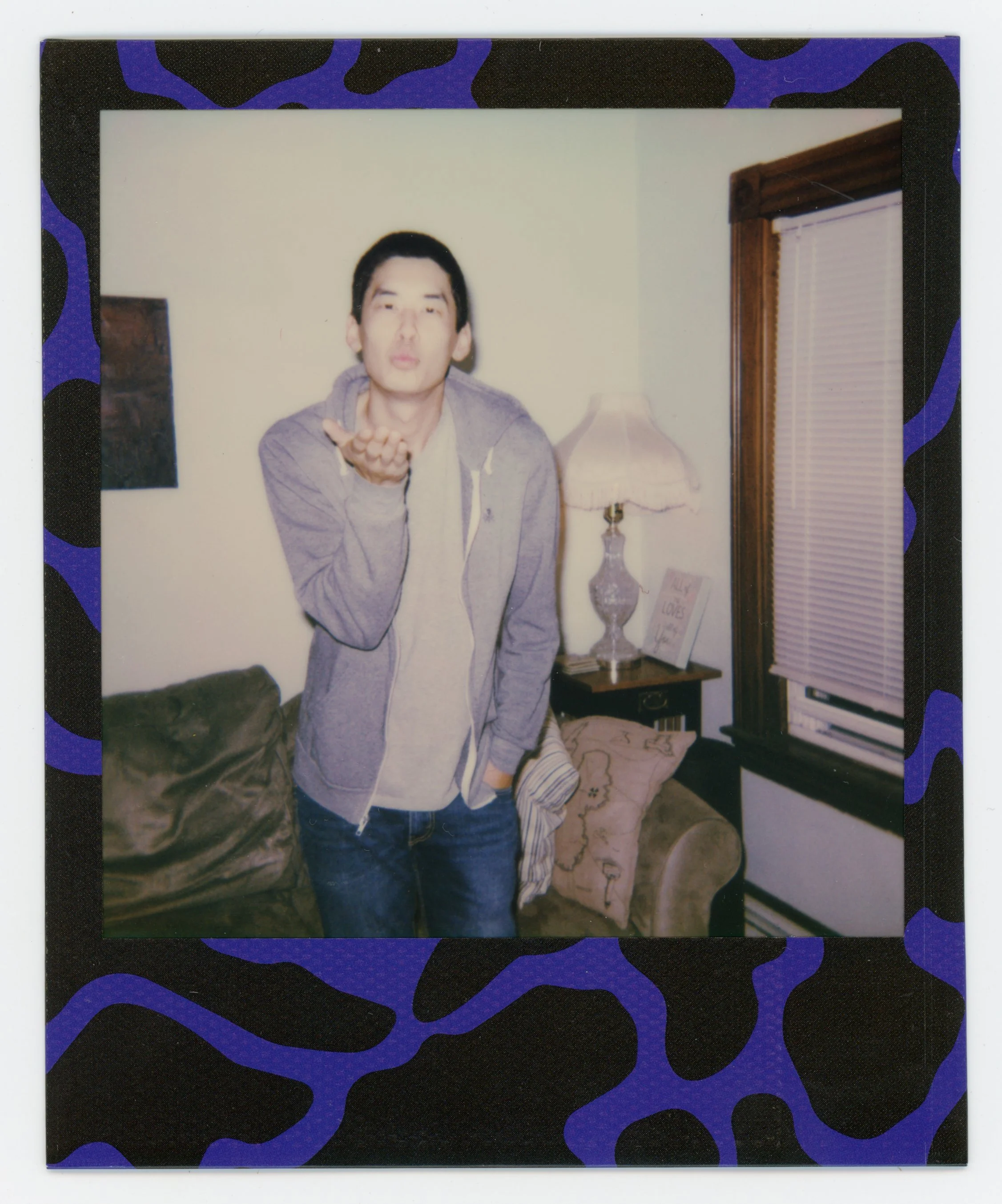 A young man with black hair, wearing a gray jacket and jeans, standing in a living room, blowing a kiss toward the camera. The room has a couch, a table lamp, and a window with blinds.