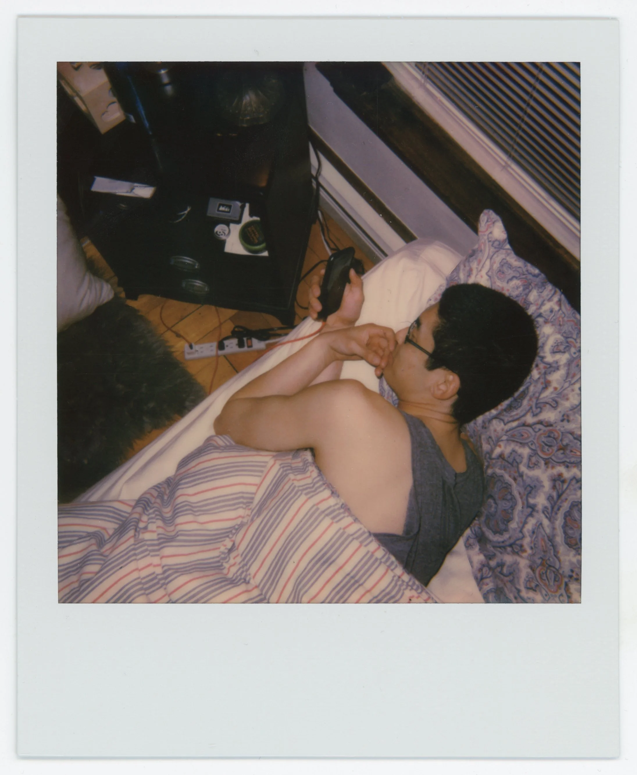 A person lying in bed, holding a remote control, with a phone near their face, wearing glasses and a sleeveless shirt, covered with striped blankets, with various items on a black shelf nearby.