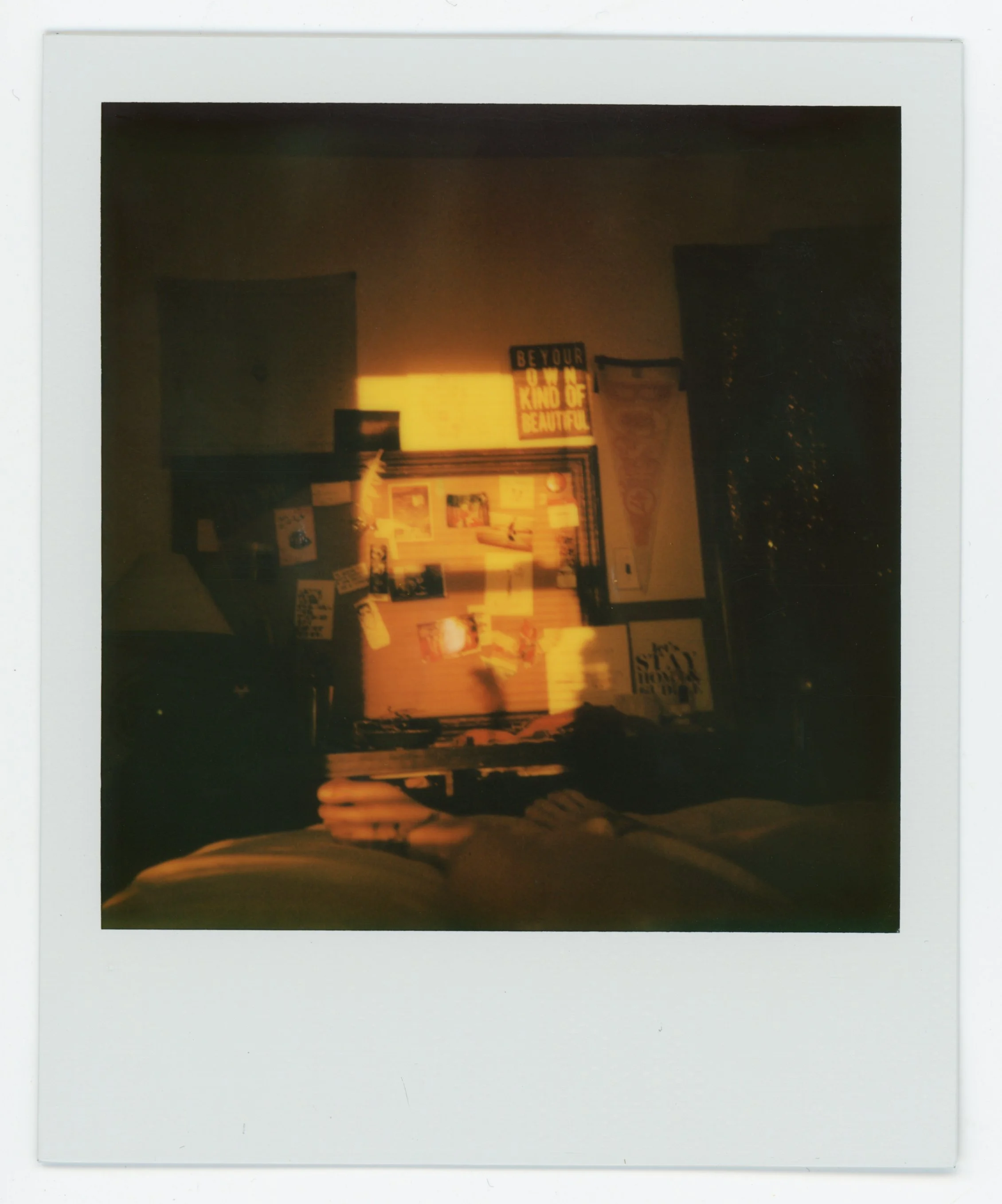 Polaroid photo showing a reflection of a person sitting indoors, with a wall decorated with posters and signs, including one that says 'BE YOUR OWN KIND OF BEAUTIFUL' and a window or door with curtains. The lighting suggests sunset or sunrise.