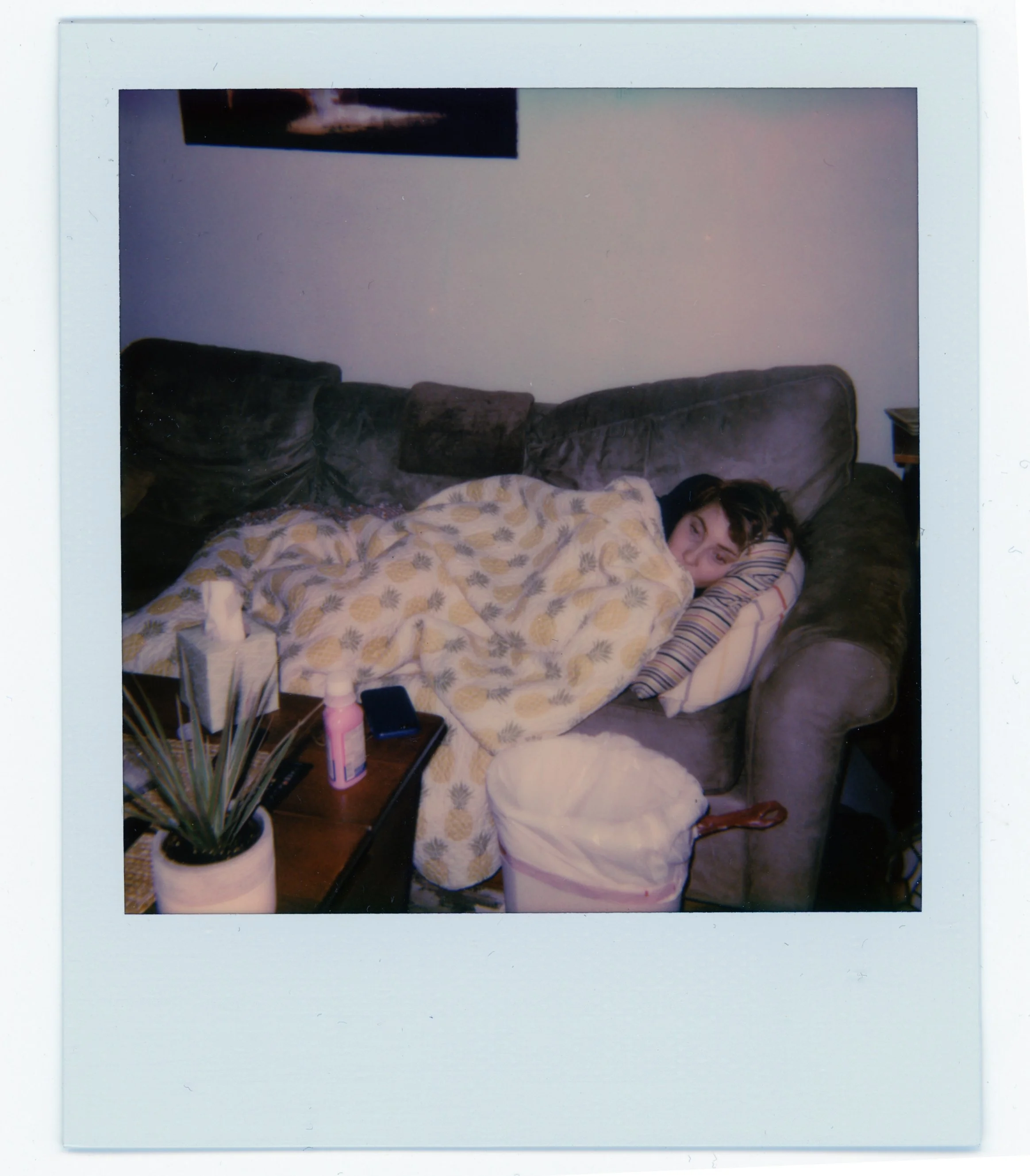 A person lying on a couch covered with a pineapple patterned blanket, resting their head on a striped pillow, with a plant, bottle, and phone on a nearby table.