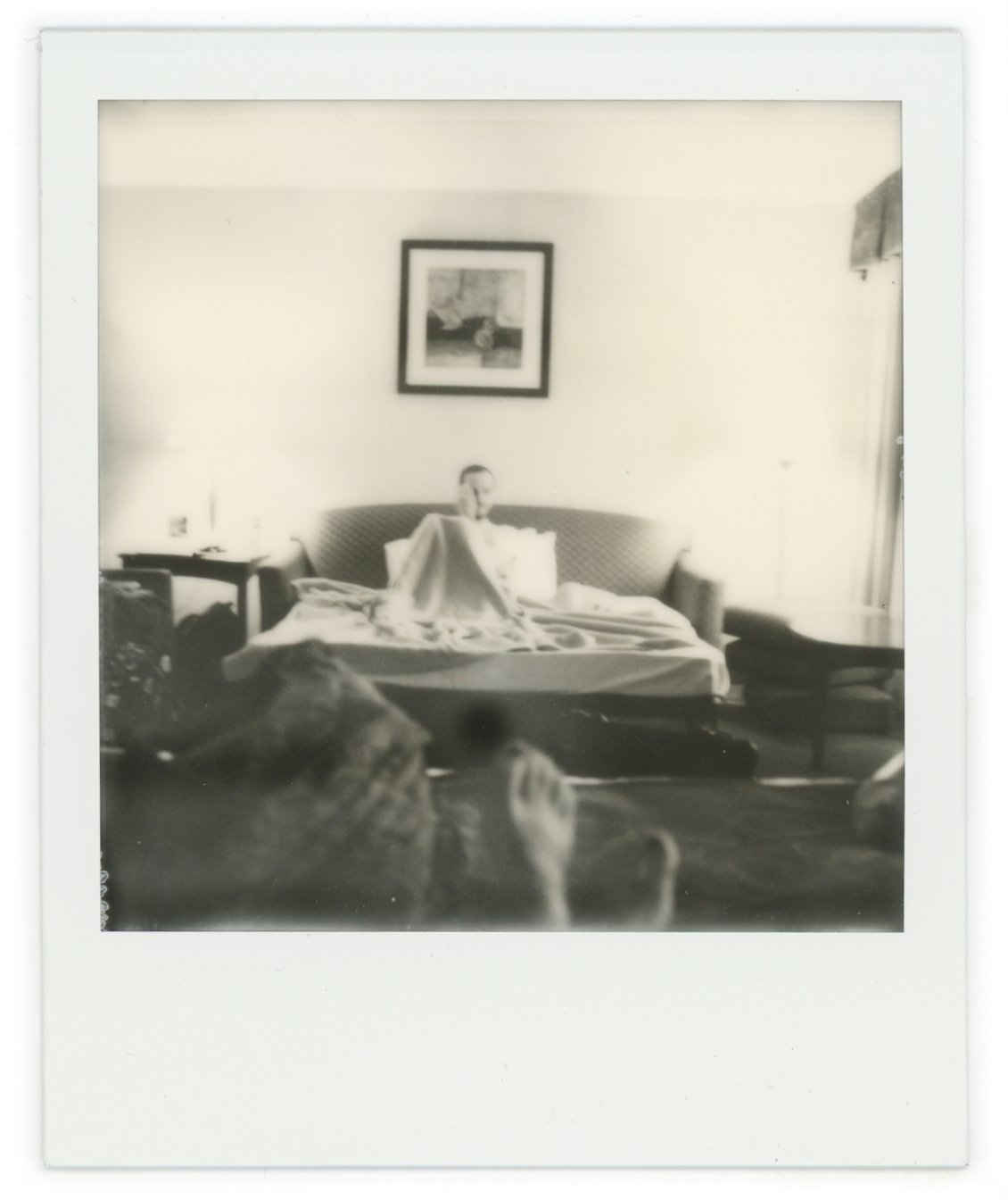 Black and white photo of a woman sitting on a bed with a headboard in a living room, with a framed picture hanging above her, and side tables on either side of the bed.