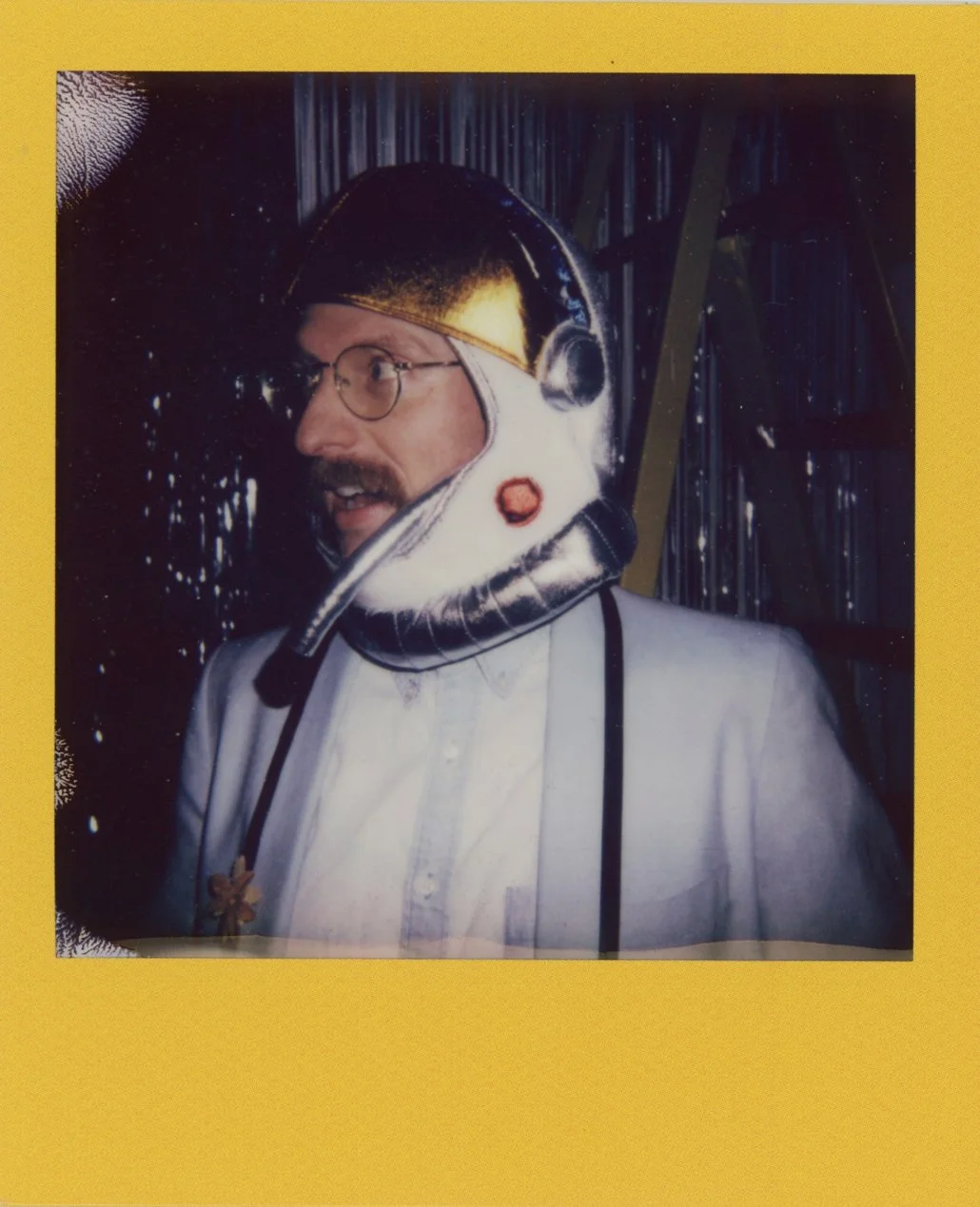 A man with glasses and a mustache wearing a space helmet, dressed in an astronaut costume, standing in front of a shiny, metallic background with yellow borders.