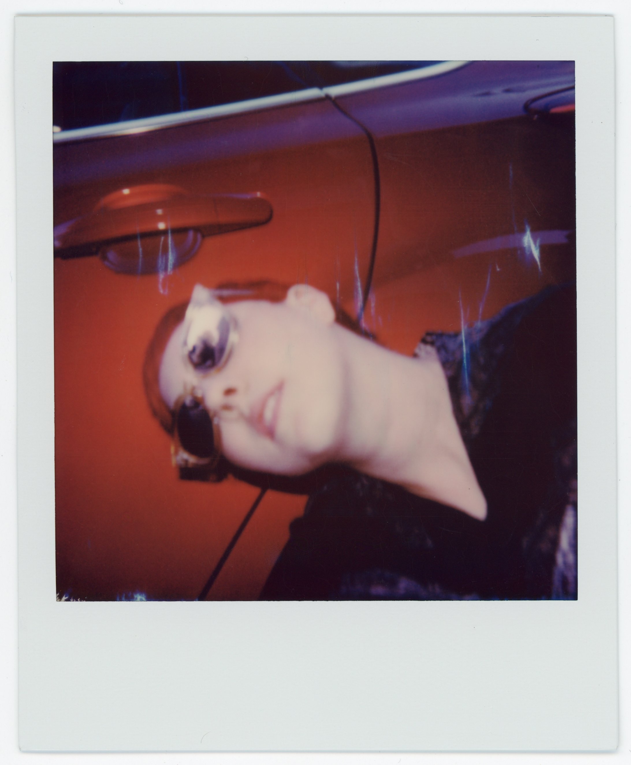 A person with short red hair wearing sunglasses lying down against a orange wall, with a person in dark clothing partially visible in the background.