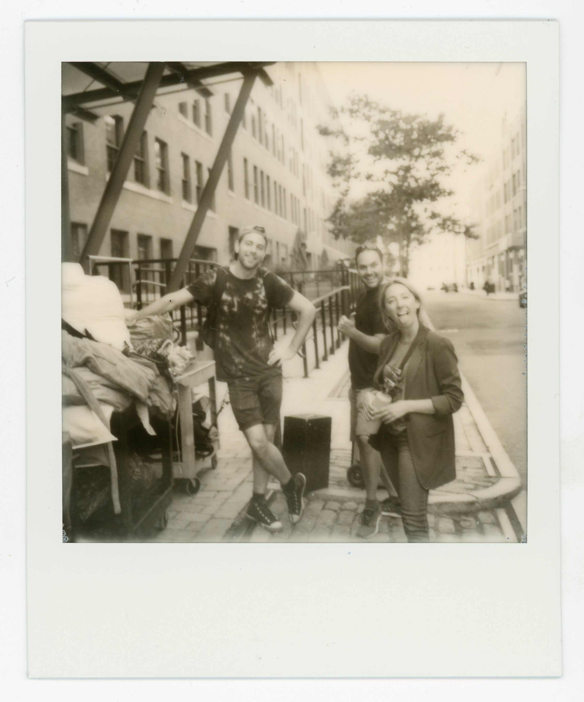 Black-and-white photo of three people smiling and posing on a city street, with buildings, trees, and parked cars in the background.