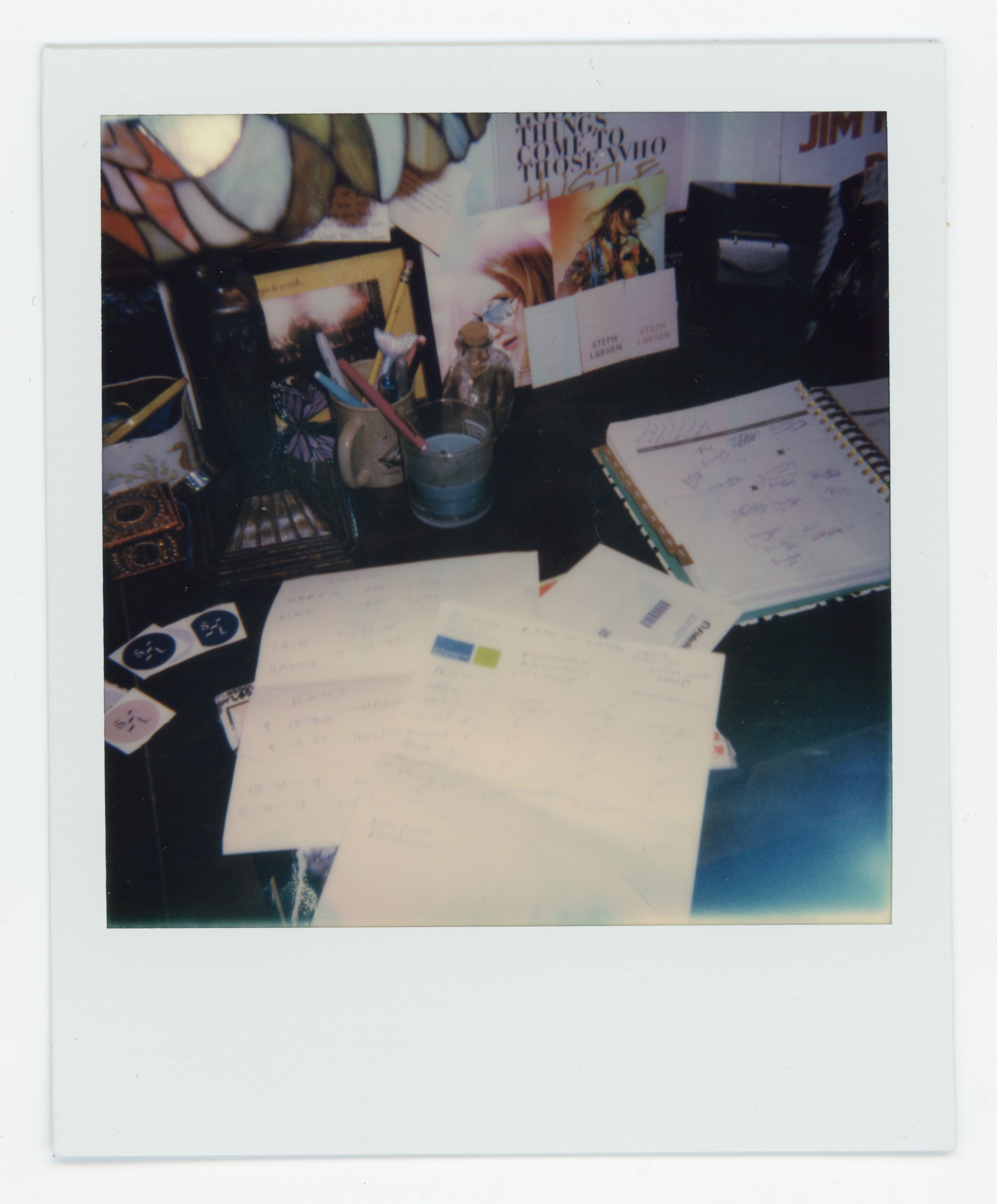 A cluttered desk with papers, notebooks, and writing supplies, including pens and markers, near books and posters.