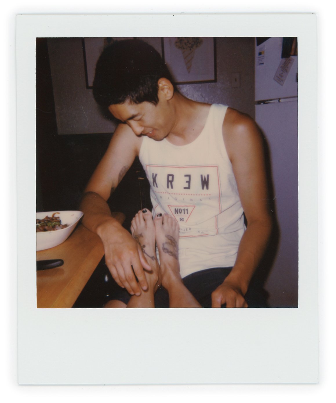 A young man sitting at a table, holding hands with a person who has tattoos on their feet and legs, with a bowl of food beside them.