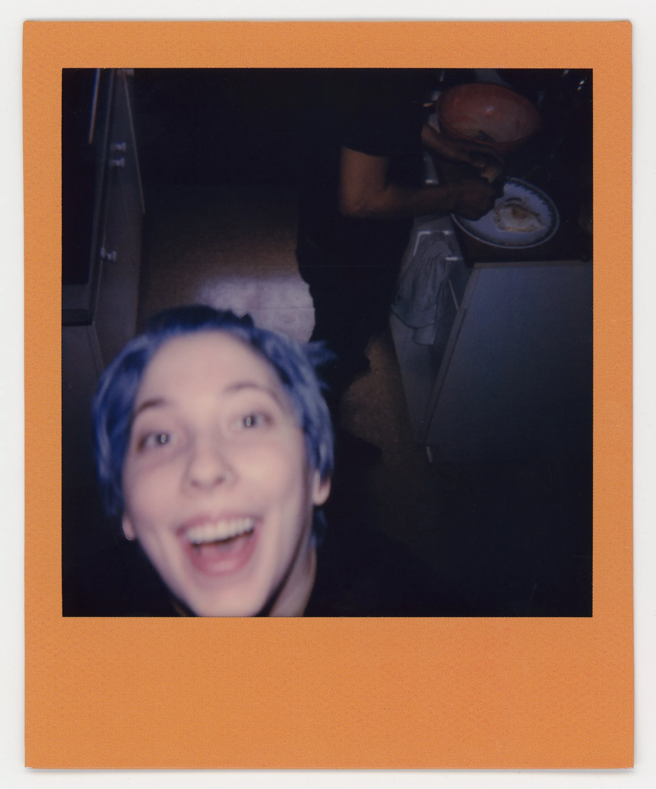 A person with purple hair smiling excitedly in the foreground, with a kitchen scene in the background where another person is standing and preparing food at the stove.