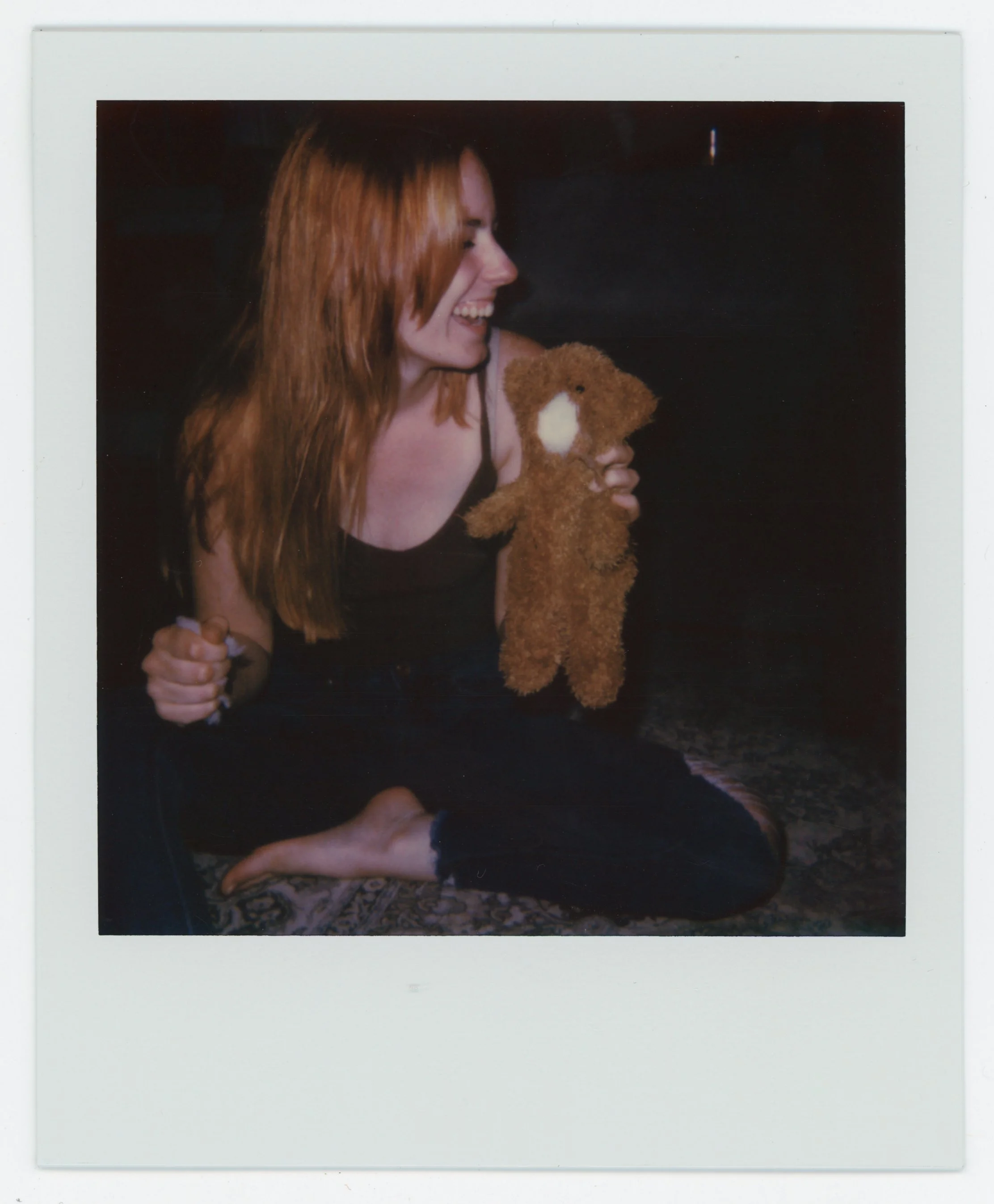 A red-haired woman sitting on the floor, holding a brown teddy bear and laughing.