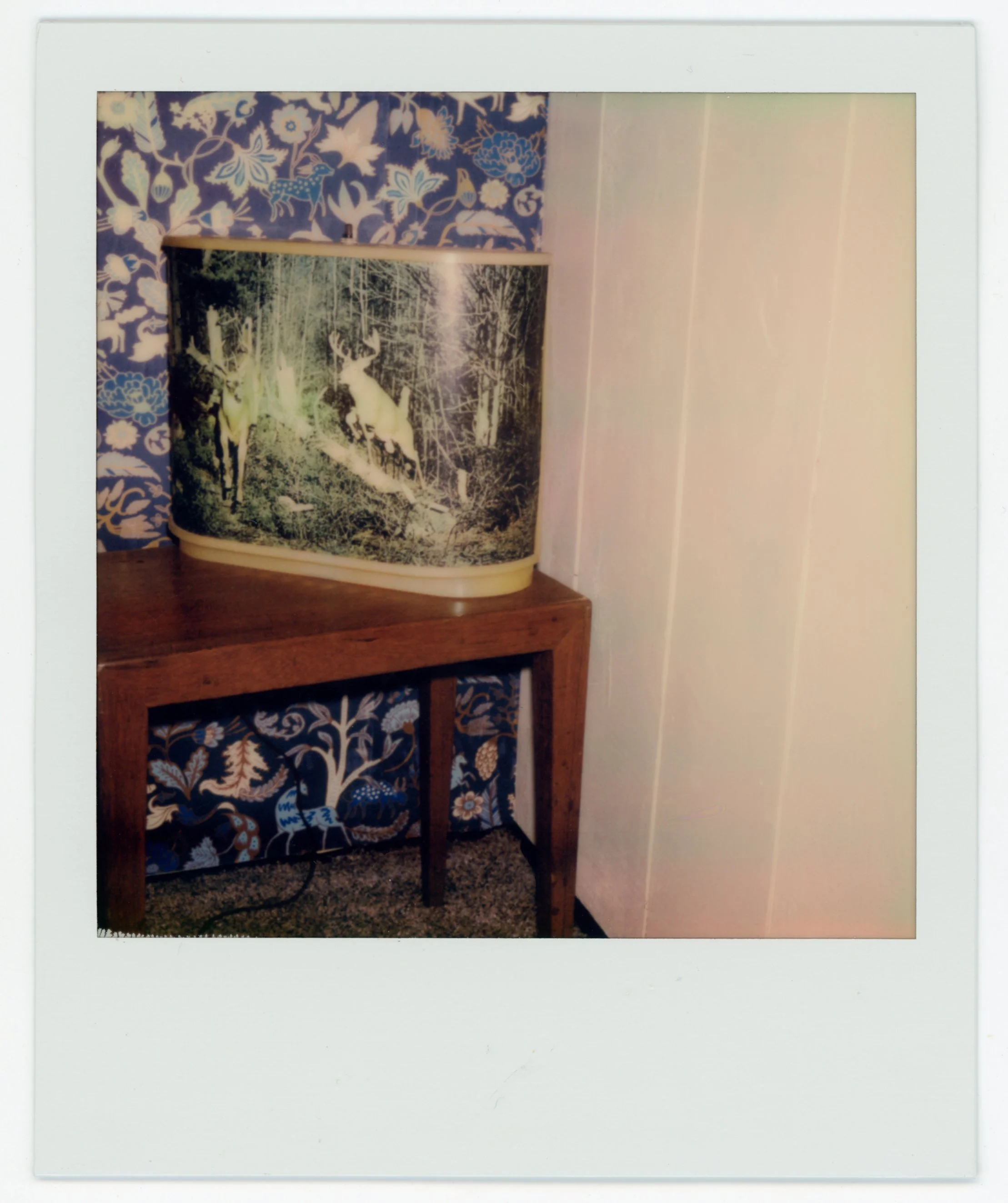 A vintage-looking lamp with a nature scene of deer, placed on a wooden table against a background of painted wallpaper and white wall paneling.