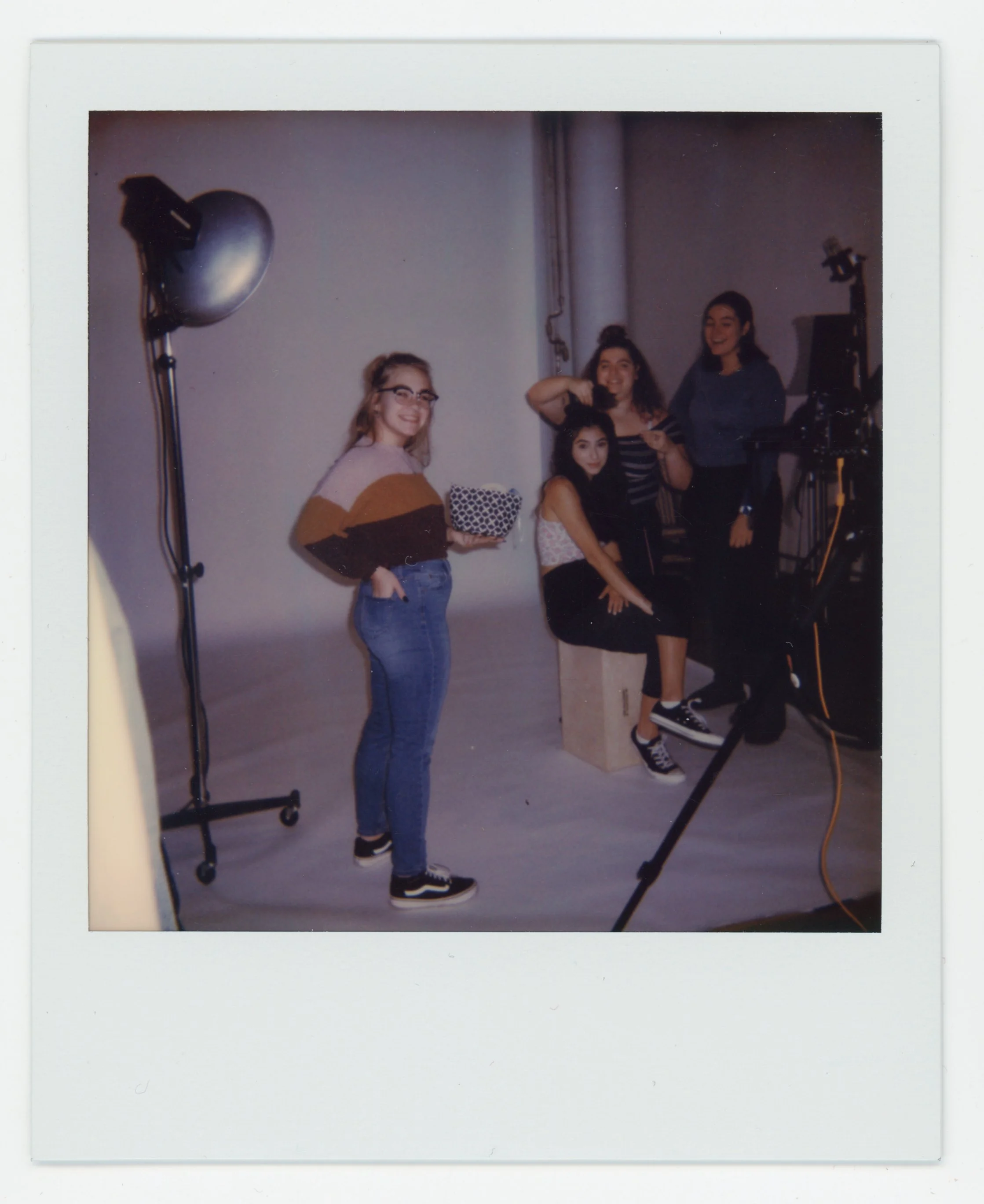 Four young women in a photography studio, two of them sitting on a box, with a camera and studio lights, one holding a gift box, and the others smiling and posing.