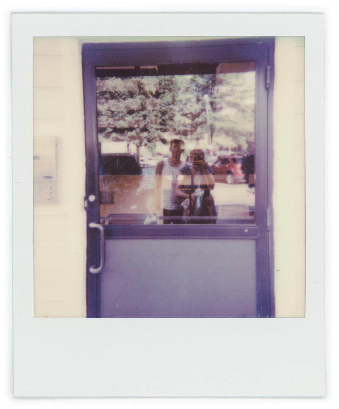 Reflection of two people taking a photo in a window, outdoors with trees and cars visible in the background.