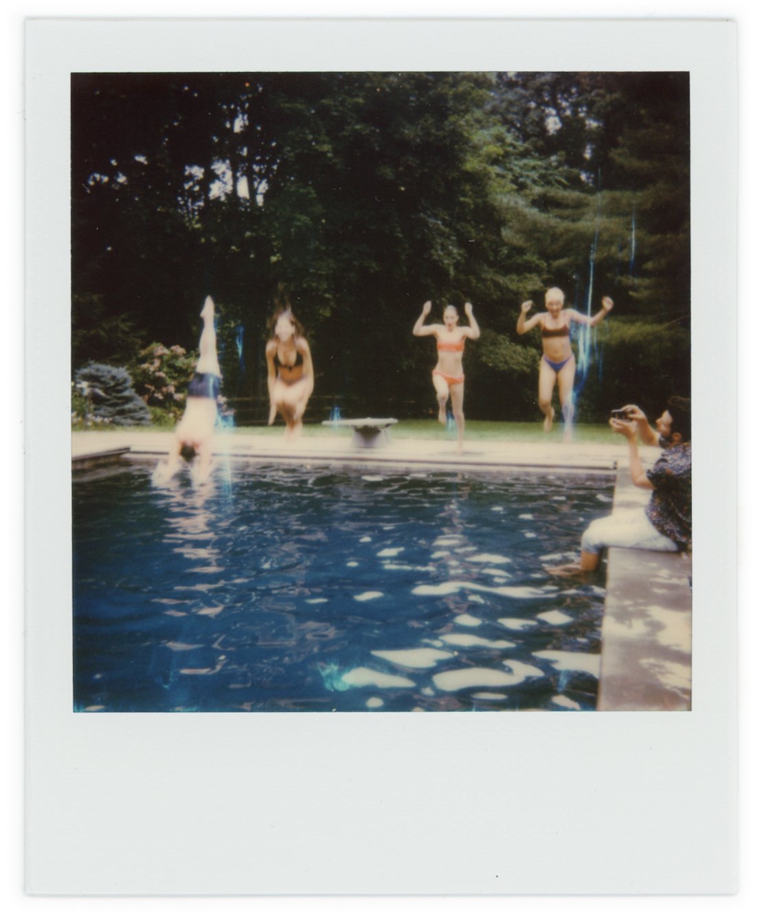 Four women jumping into a pool from a ledge, with trees in the background, and a woman sitting on the edge taking a photo.