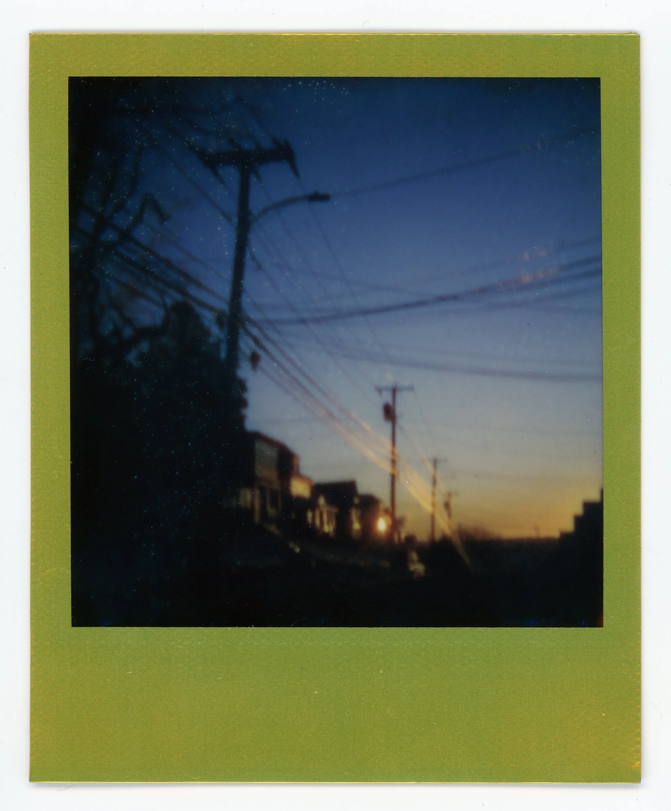 Blurred photo of a neighborhood at dusk with power lines and a house silhouette.