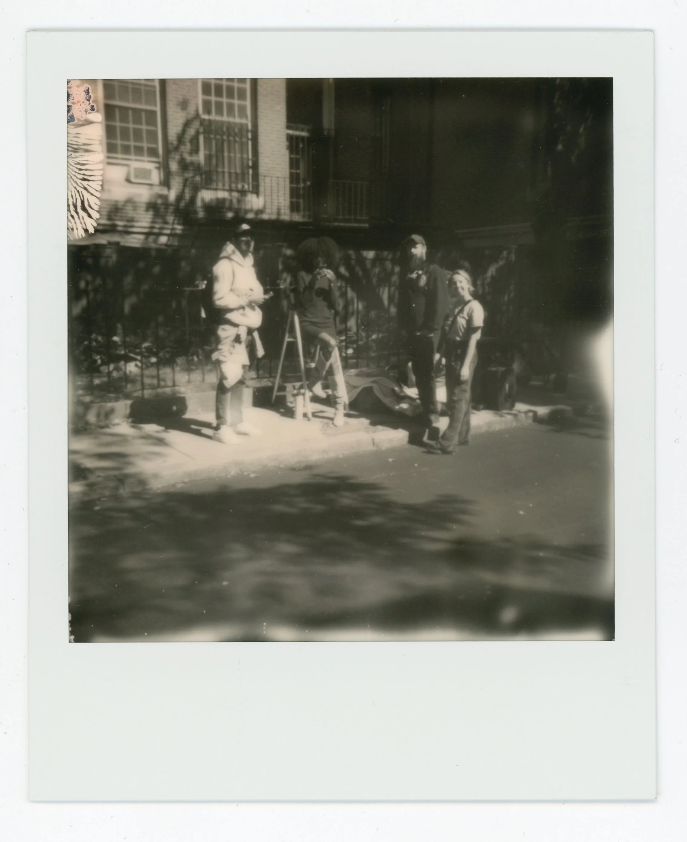 Black and white photo of four people standing on a sidewalk next to a fence and apartment building, engaged in conversation. One person is using a tripod or easel.