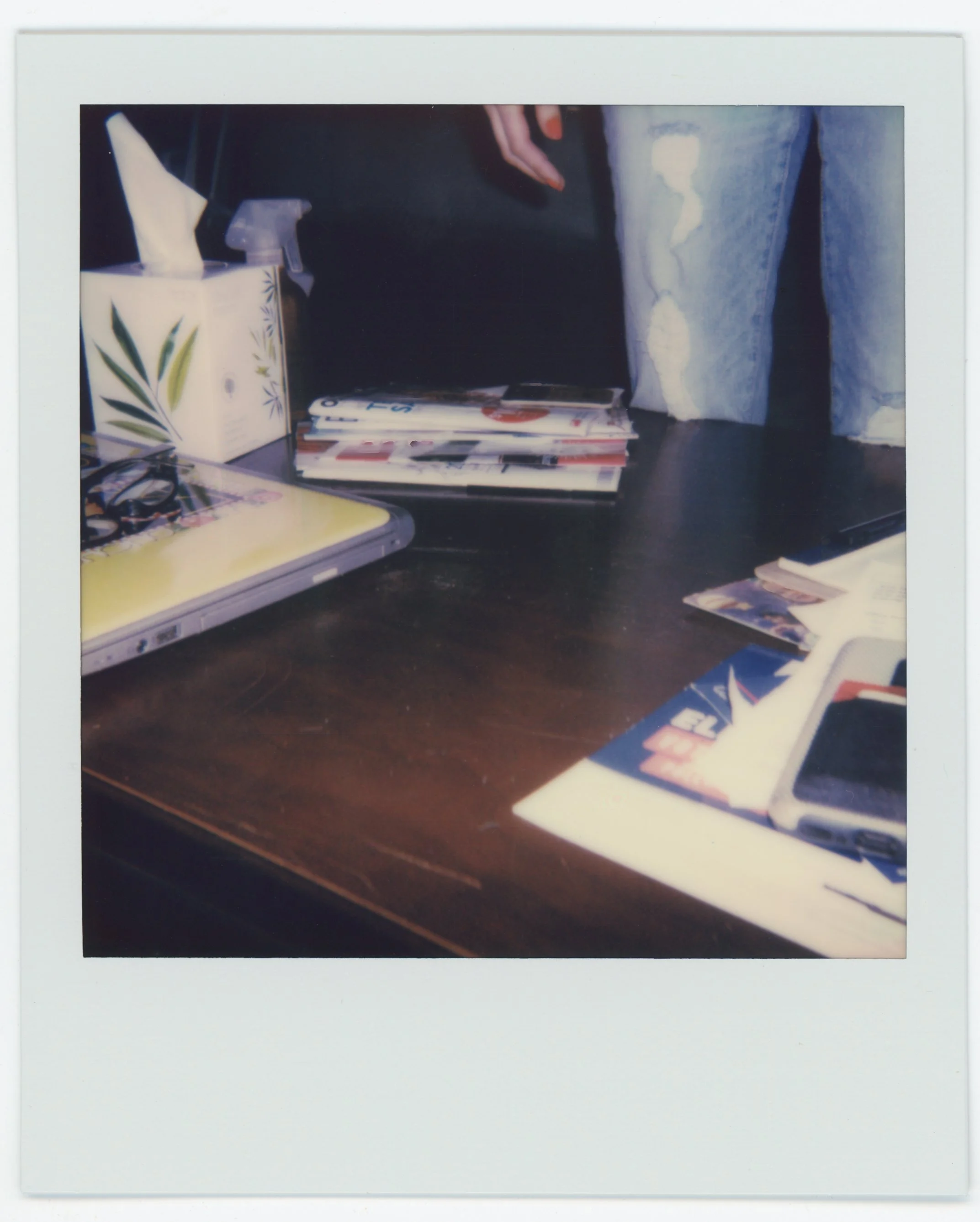 A cluttered wooden desk with an open laptop, magazines, and a phone, with a tissue box and hand sanitizer in the background.
