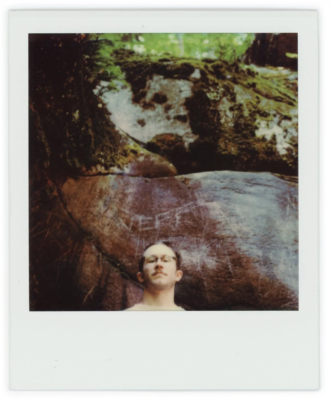 A person with glasses standing in front of a large rock with a face-like pattern in a forest setting.