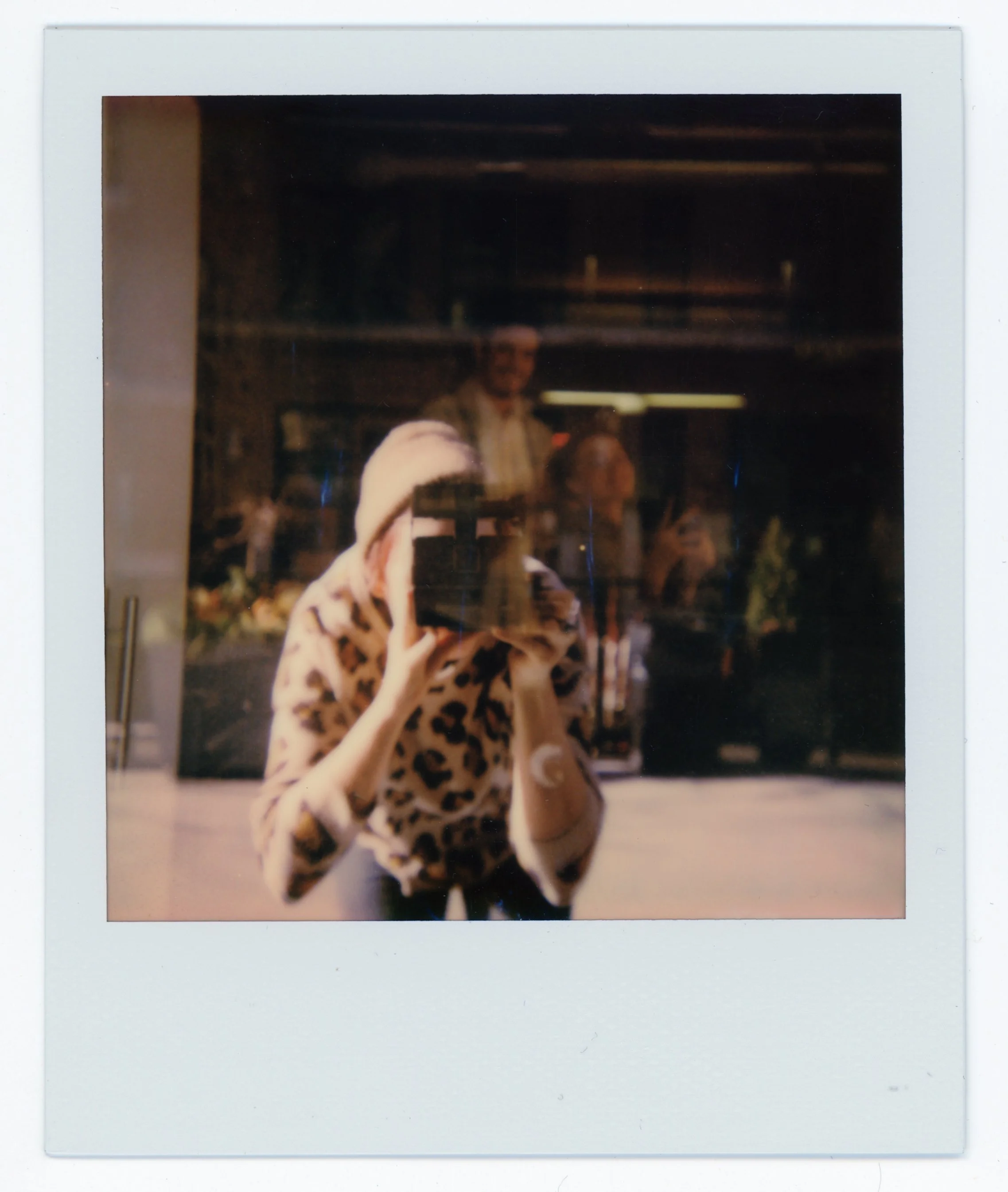 A person taking a photo with a camera through a mirror, wearing a leopard print shirt and a beanie, with other people reflected in the background.