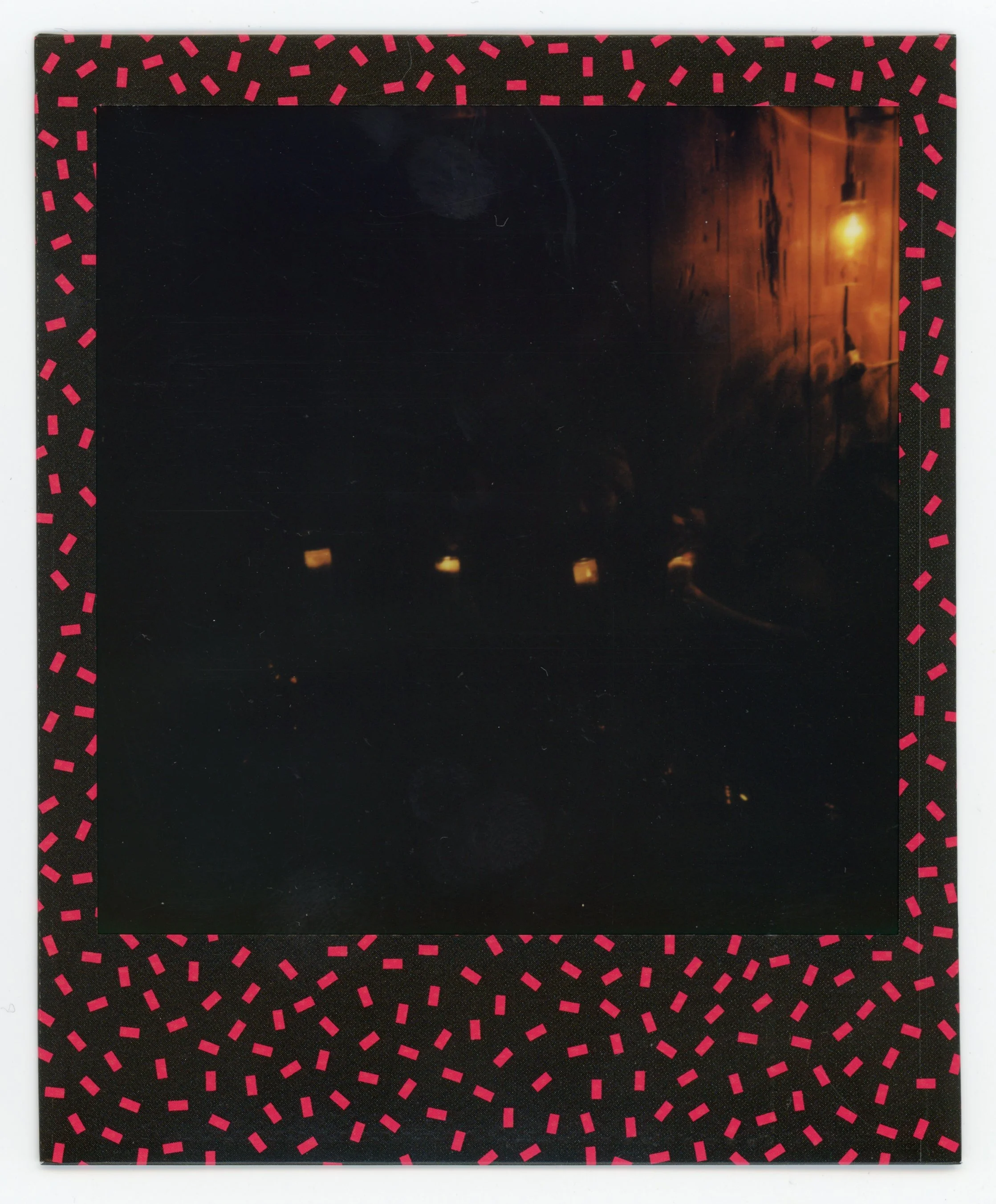 A Polaroid photograph with a black frame decorated with pink dashed lines. The photo is dark and appears to be a nighttime scene with faint lights and an orange glow on the right side.