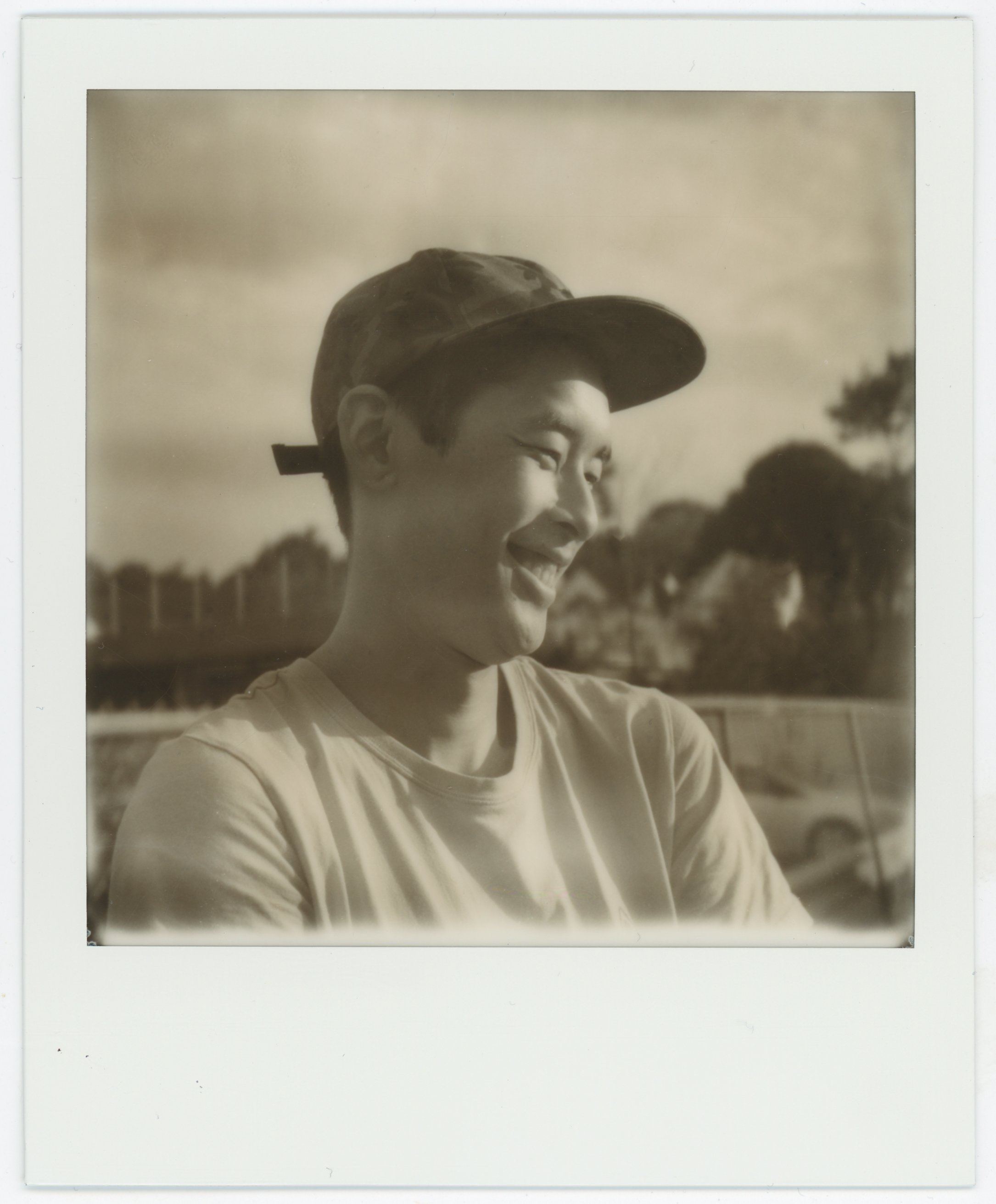 A young person wearing a cap, smiling and looking to the side outdoors with trees and a vehicle in the background.