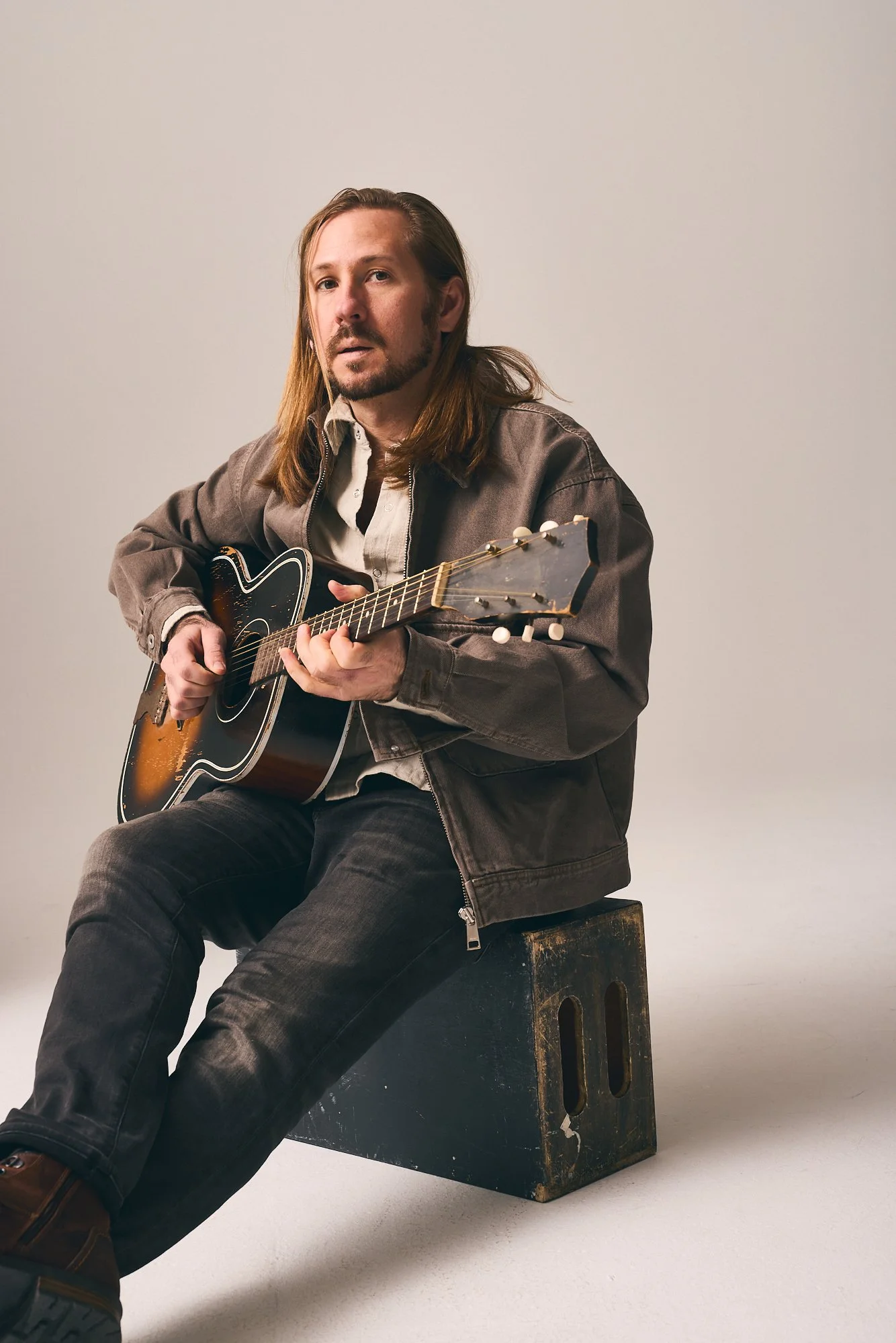 A man with shoulder-length brown hair and a beard, wearing a brown jacket and beige shirt, is sitting on a black box and playing an acoustic guitar against a plain light-colored background.