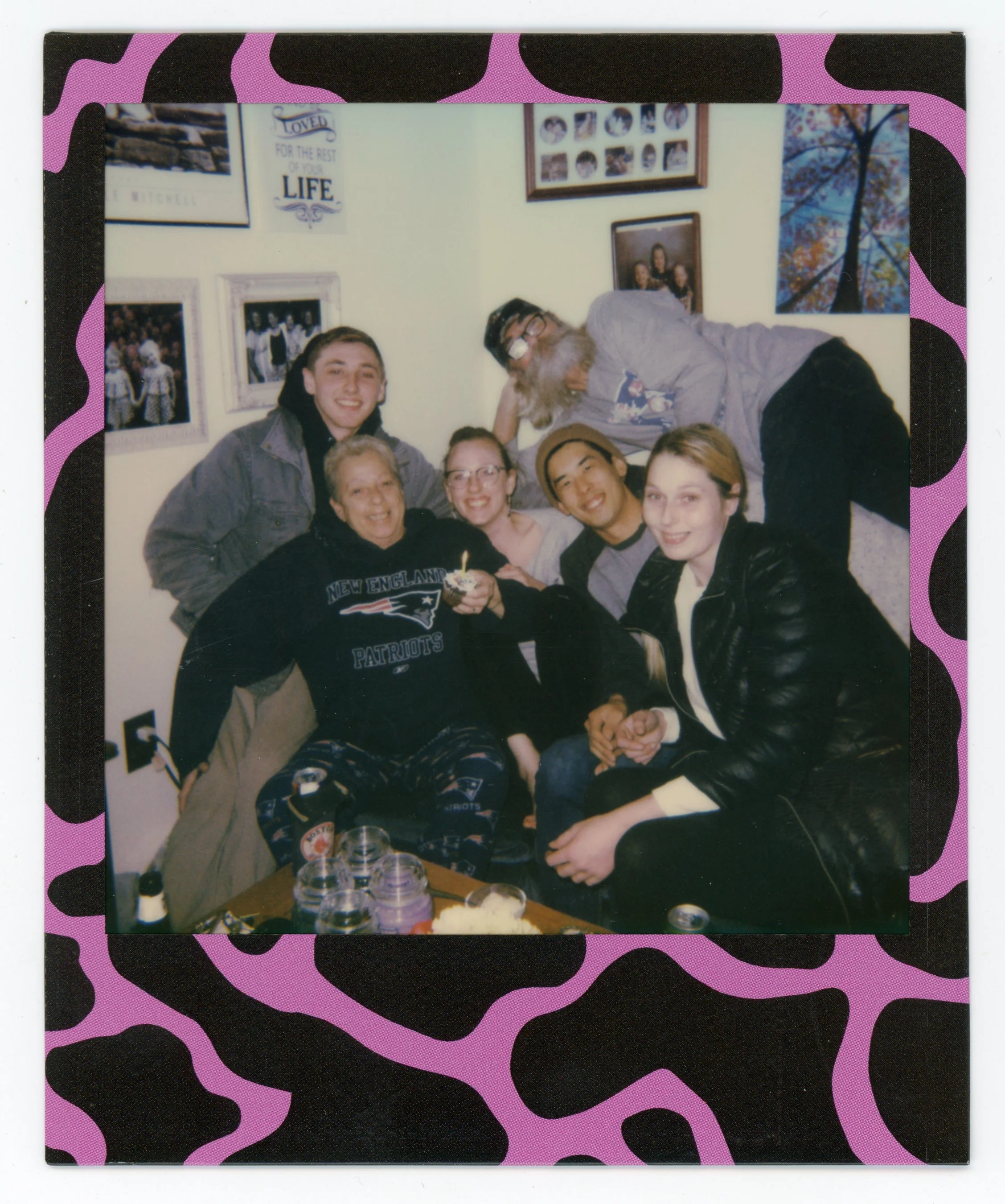 Group of six people celebrating a birthday, sitting together and smiling, with a woman holding a cupcake with a lit candle. The scene is in a cozy room with family photos on the wall and snacks on a table in front.