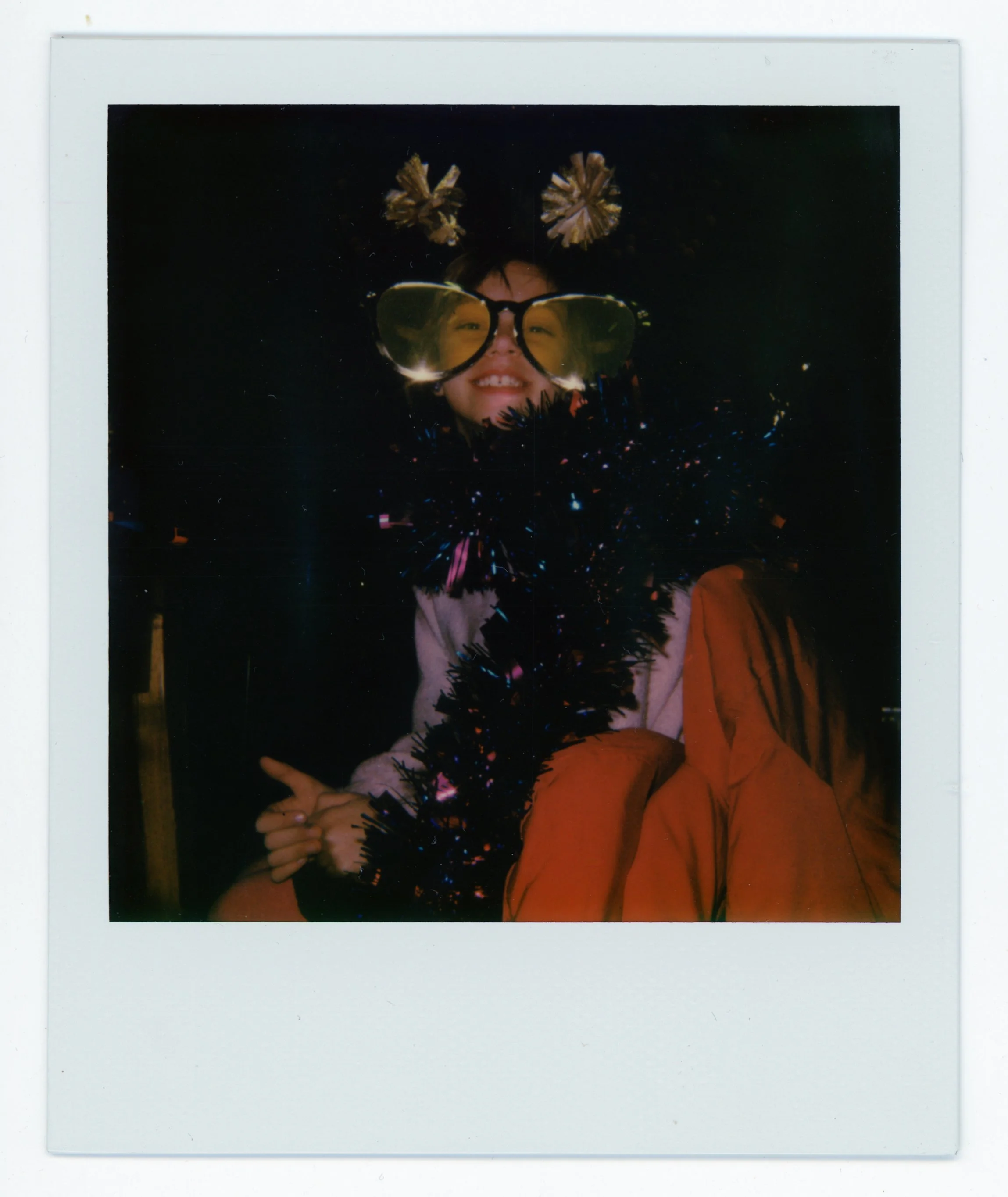 A young girl wearing large yellow sunglasses, a festive headband with gold pom-poms, and colorful tinsel garland around her neck, smiling at the camera, with a dark background.