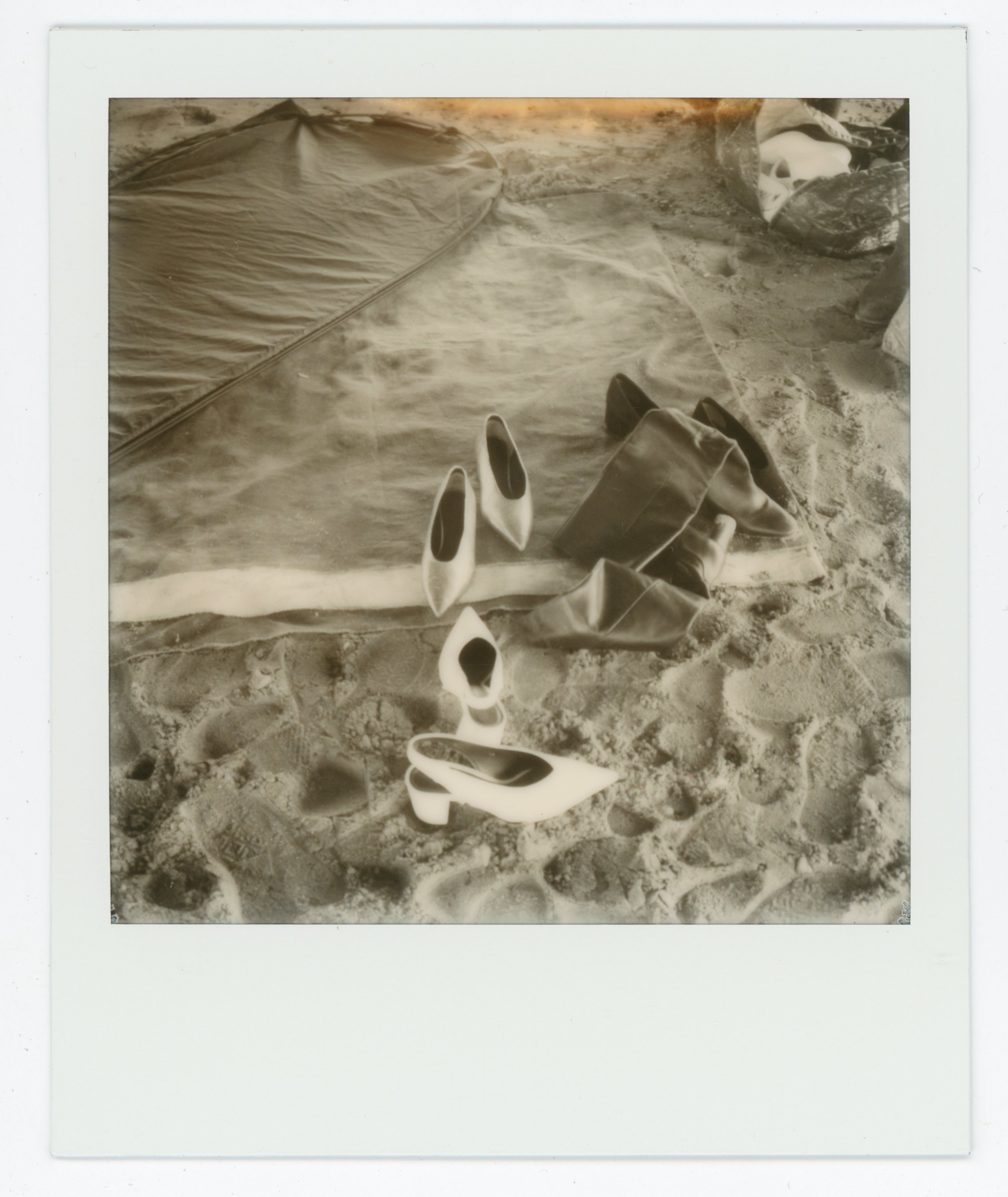 Collection of high heel shoes, a bag, and other items on sandy ground near a tent, depicted in a sepia-toned photograph.