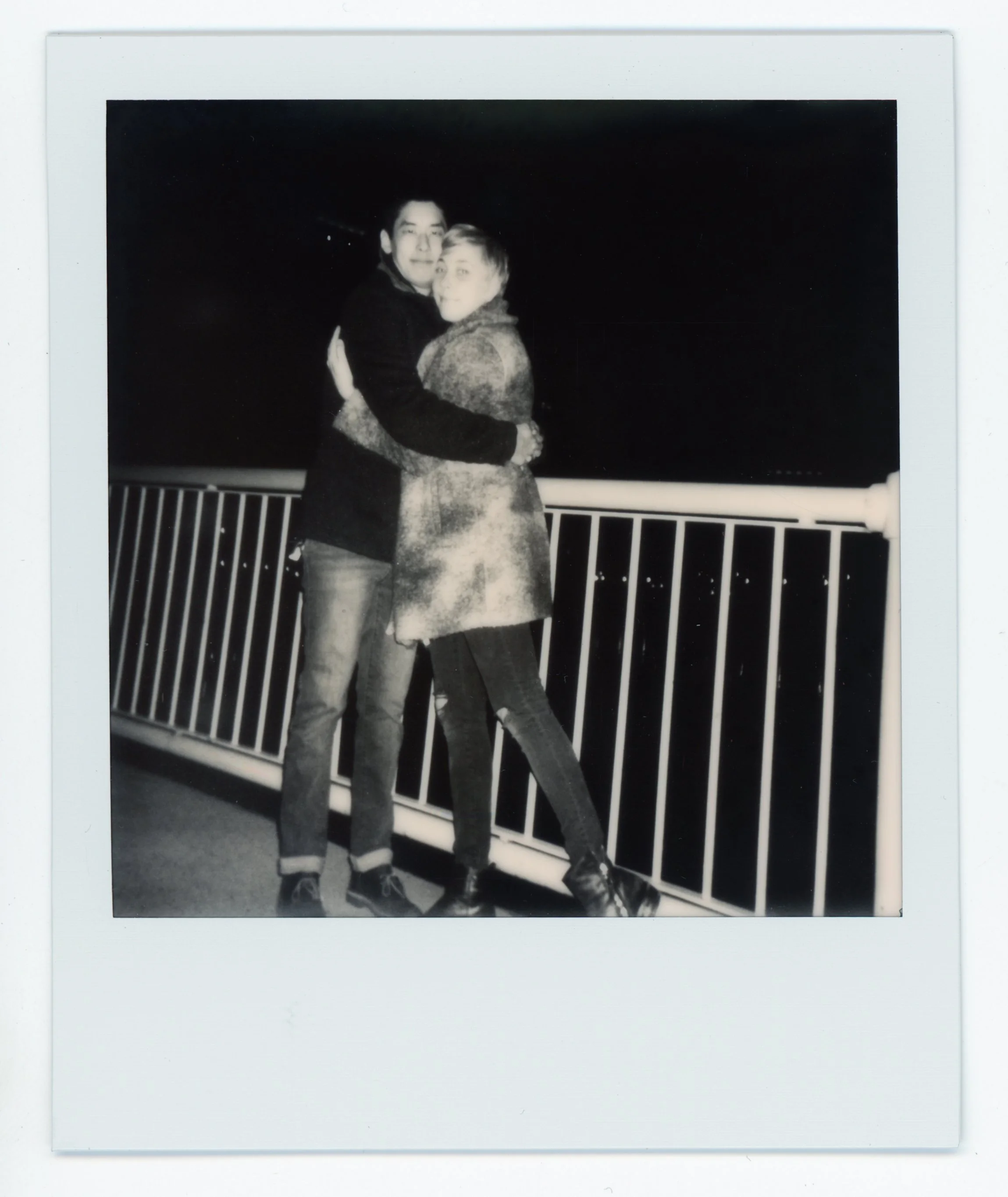 Two people hugging at night on a balcony with a railing, in black and white.