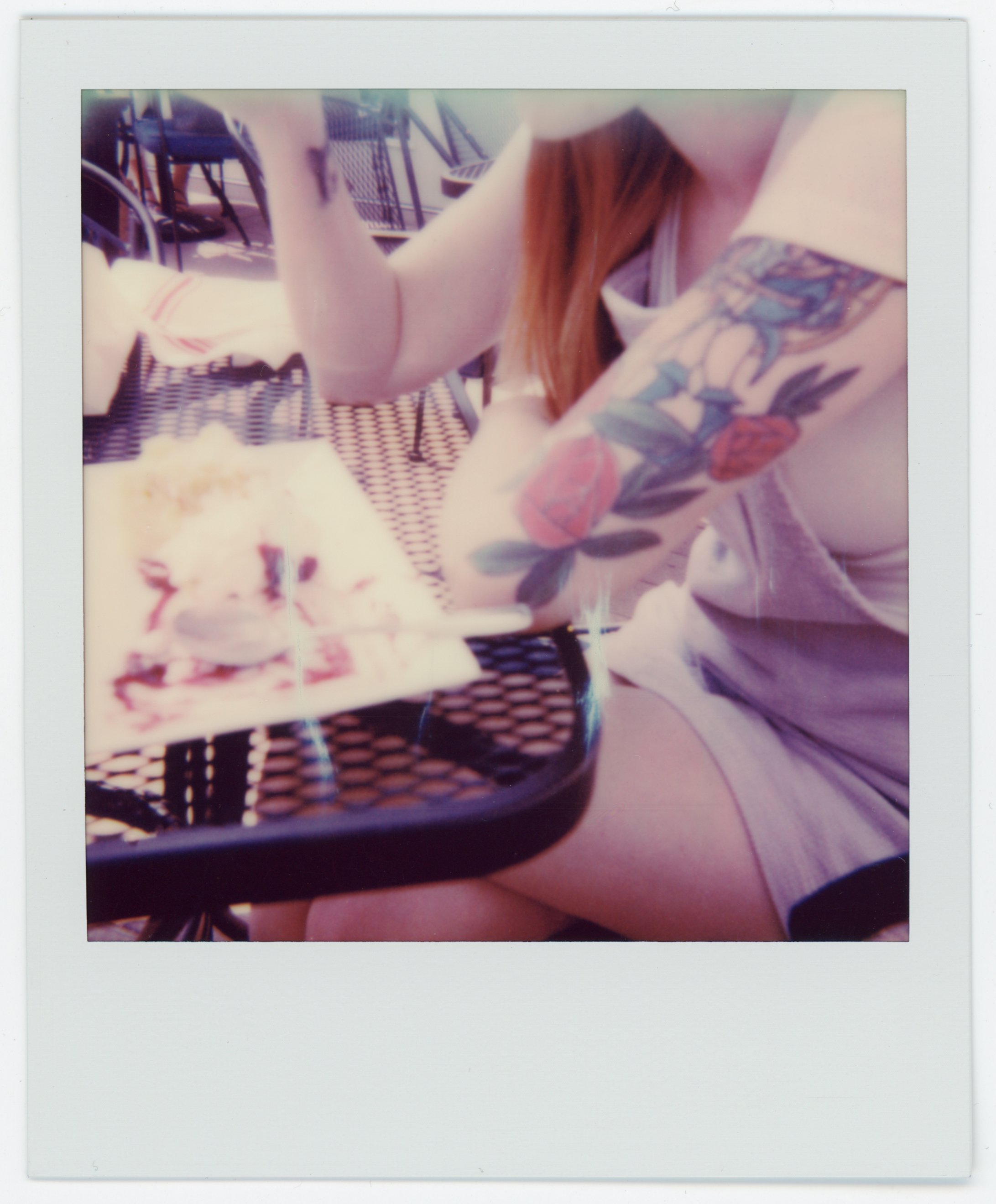 Person with a colorful tattoo on their arm, holding a fork, sitting at an outdoor table with food and a birthday candle in the dessert.