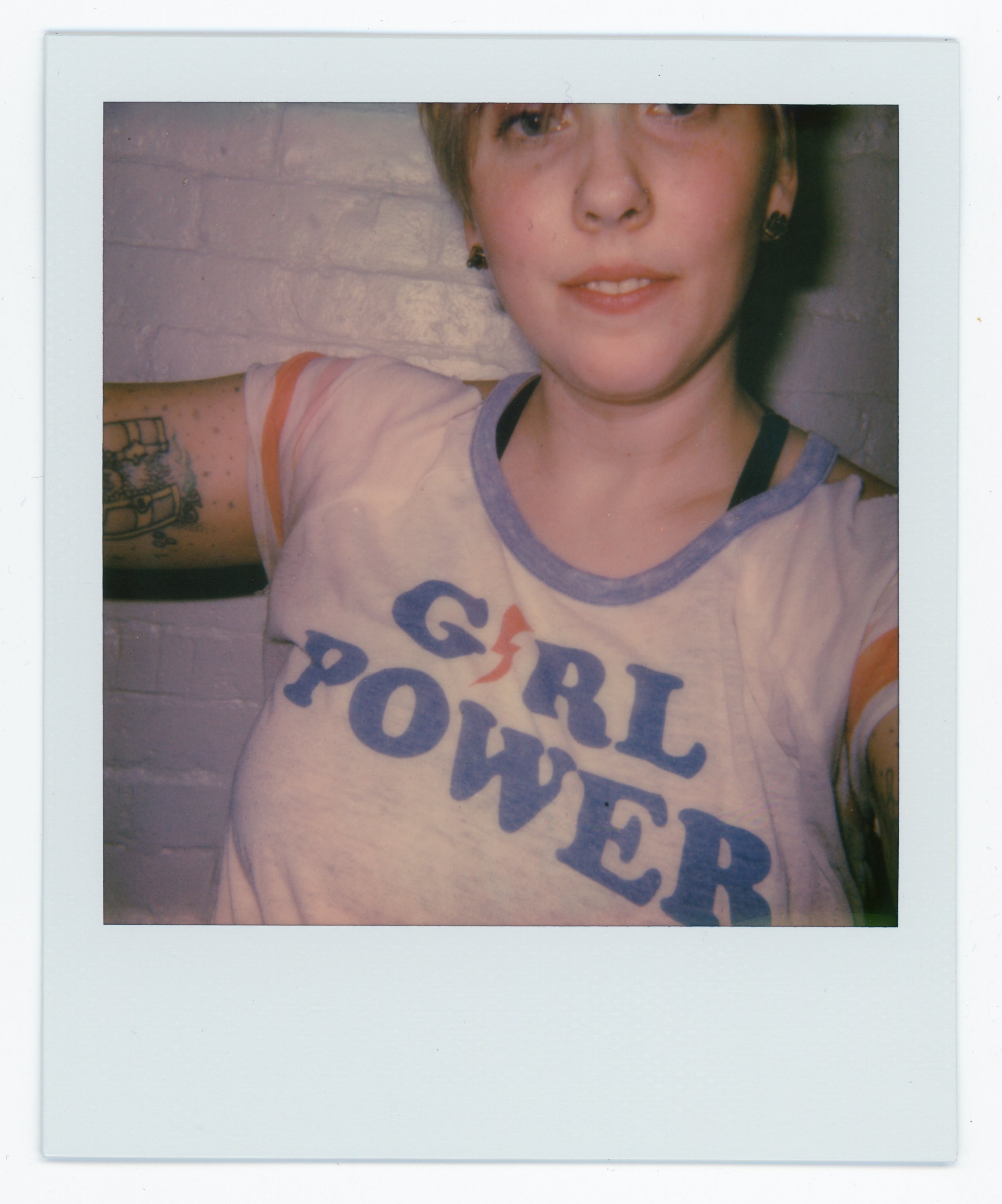 A person with short hair taking a selfie, wearing a white t-shirt that says 'GIRL POWER' in blue and red letters, with tattoos on their left arm and earrings, standing against a brick wall.