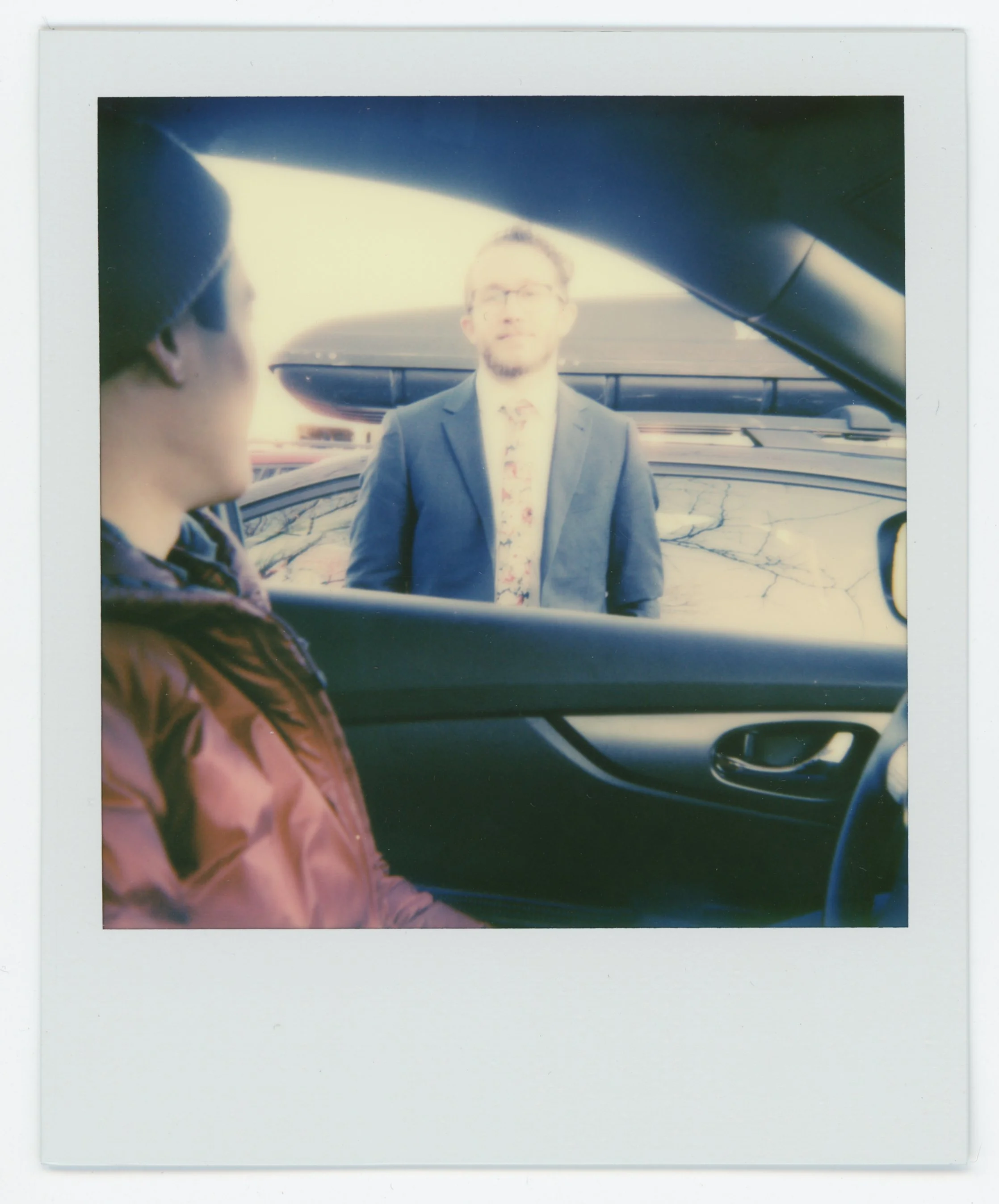 A person sitting inside a car looking at another person standing outside, viewed through the car window. The person inside is wearing a purple jacket, and the person outside is dressed in a suit with a floral shirt.