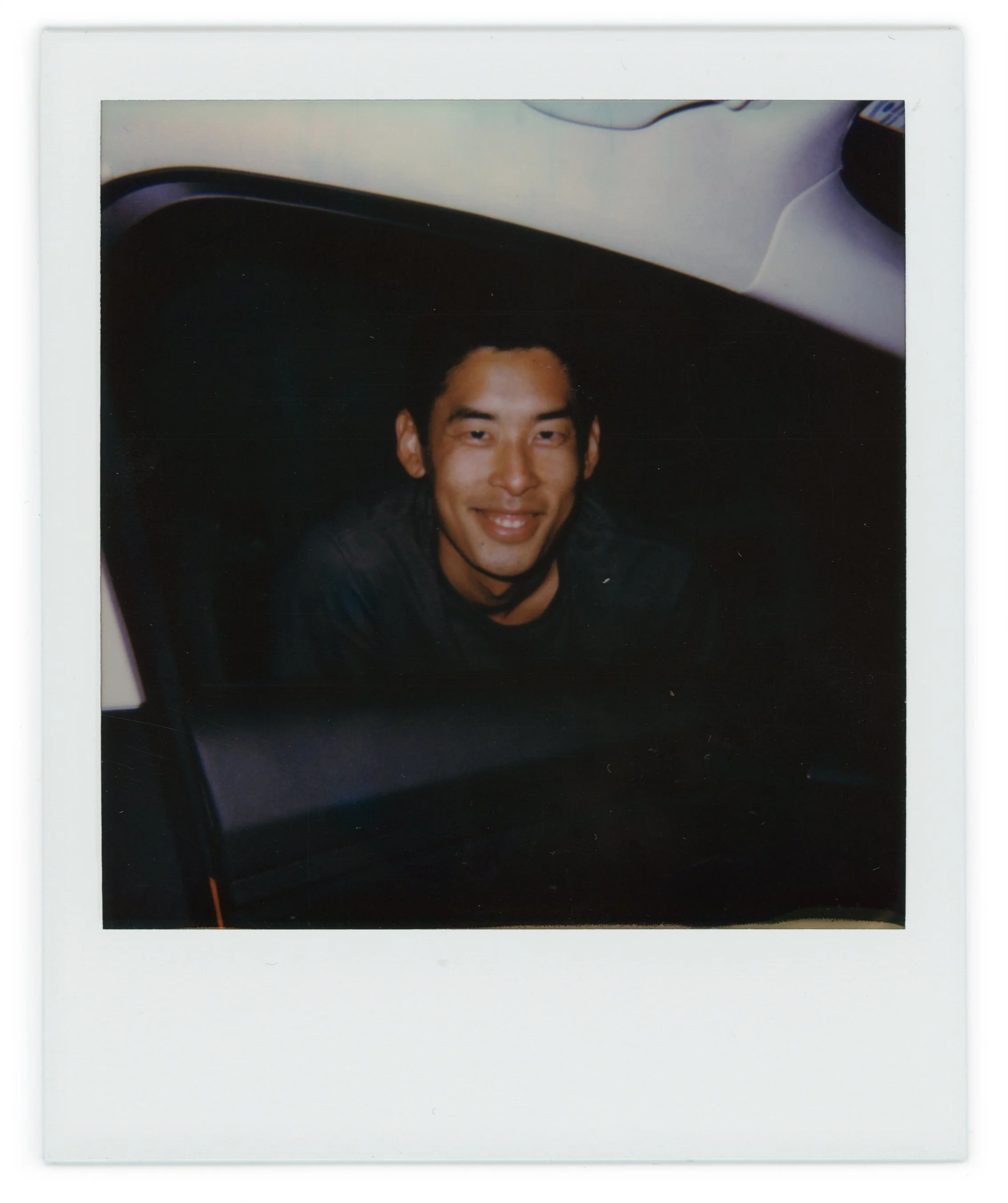 A young man with dark hair and a big smile, sitting inside a car with the window down.